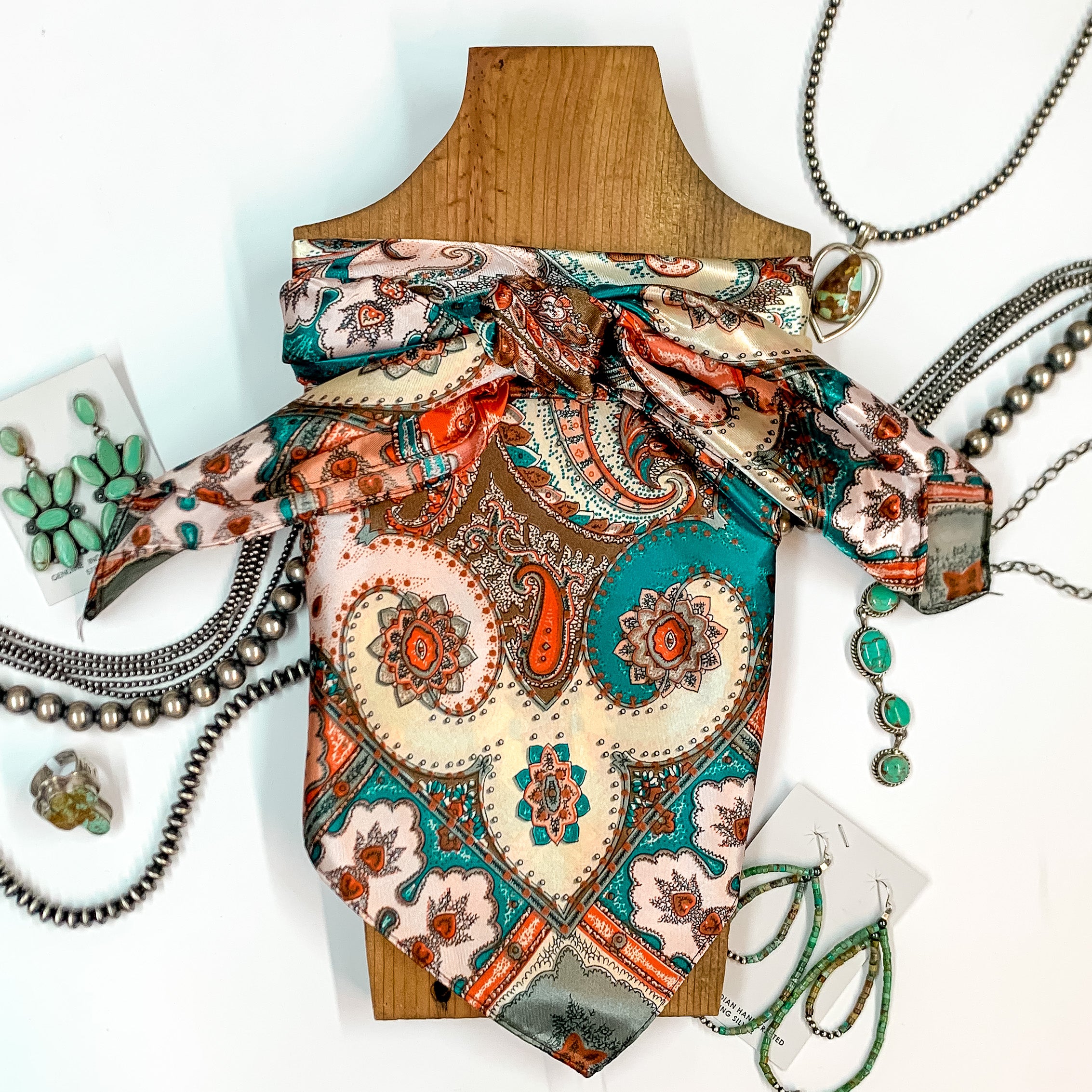 Patterned scarf in Greeley. Scarf is tied around a wooden piece. The scarf and piece of wood is pictured on a white background with Navajo jewelry spread out around it.