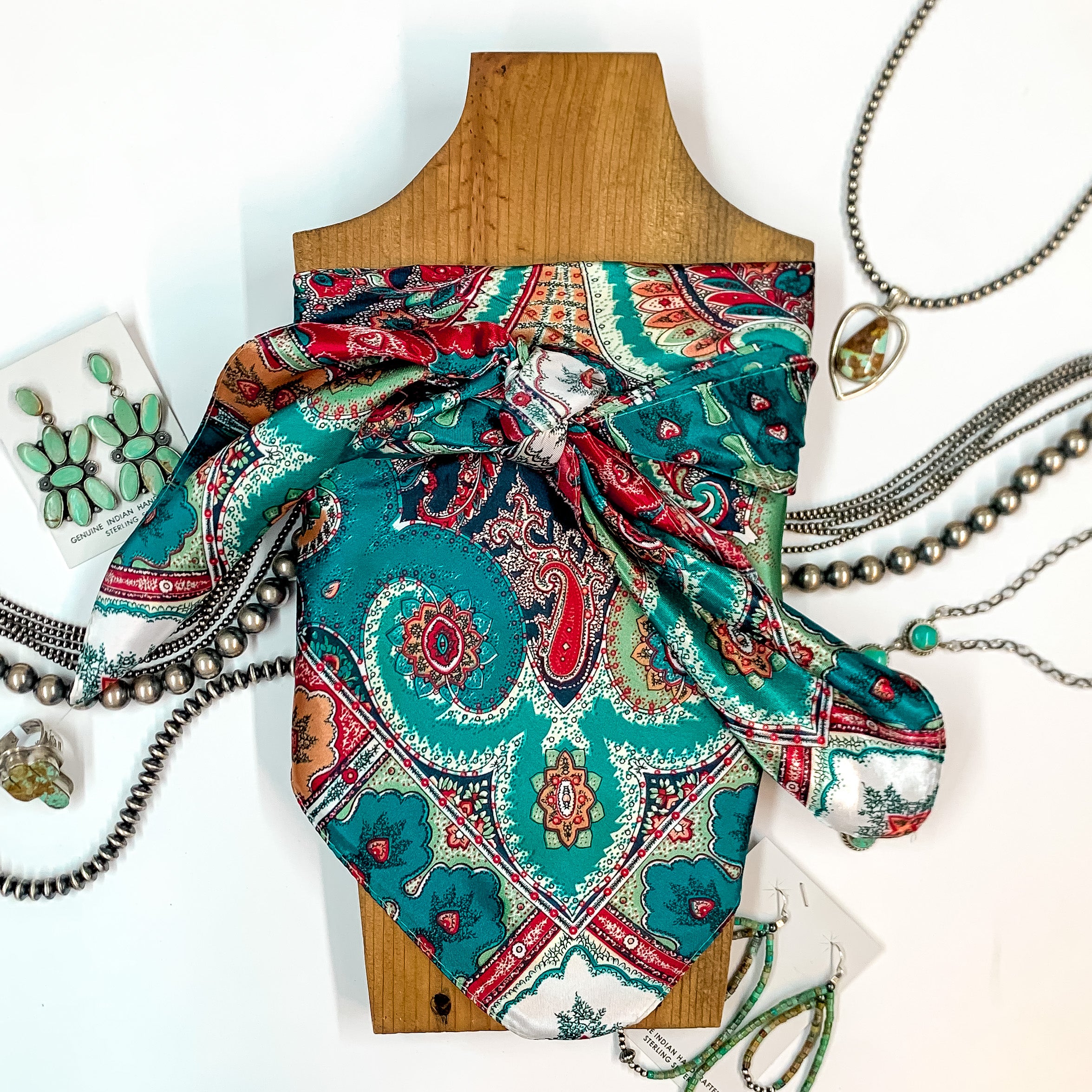 Patterned scarf in Sheridan. Scarf is tied around a wooden piece. The scarf and piece of wood is pictured on a white background with Navajo jewelry spread out around it.