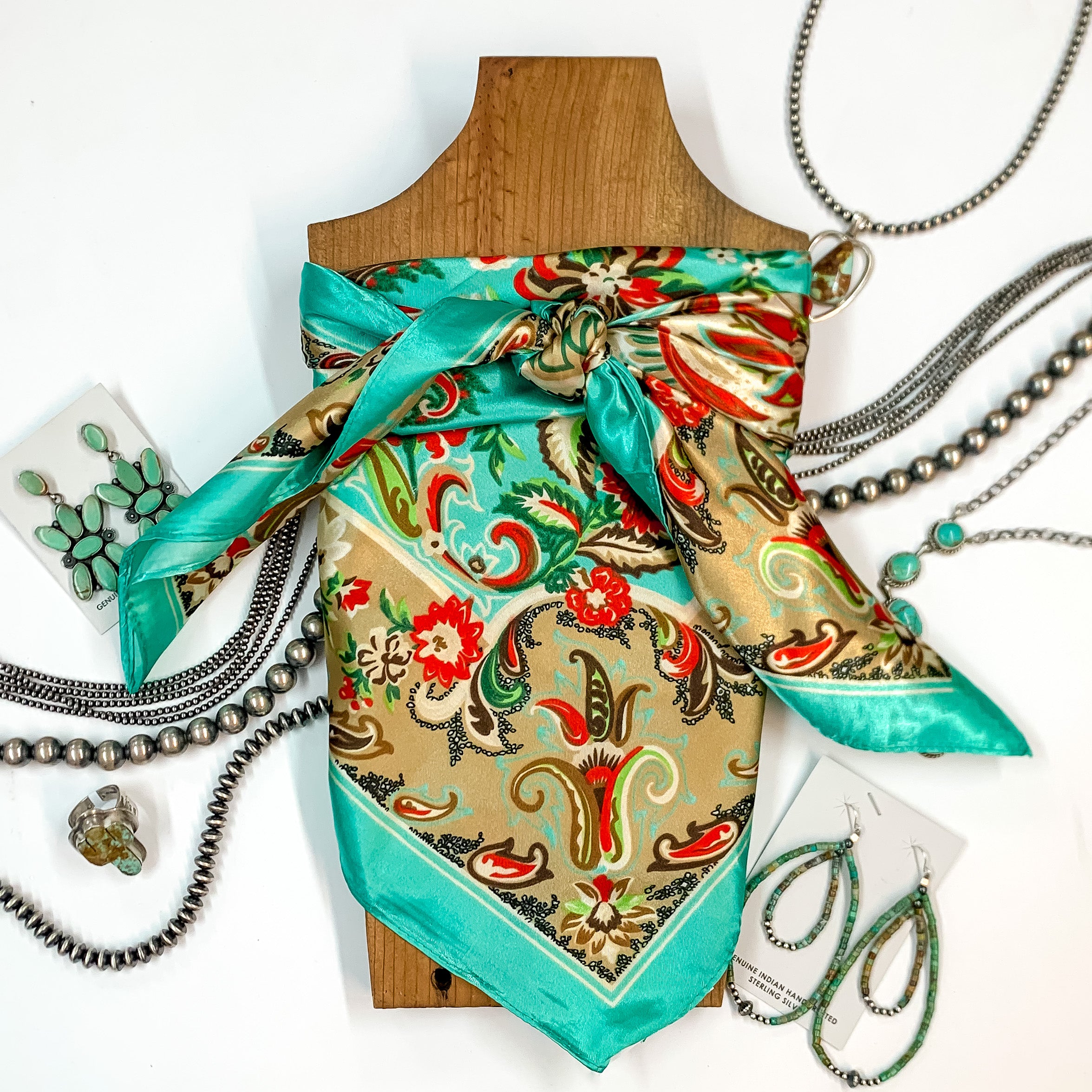 Patterned scarf in Taos Valley. Scarf is tied around a wooden piece. The scarf and piece of wood is pictured on a white background with Navajo jewelry spread out around it.