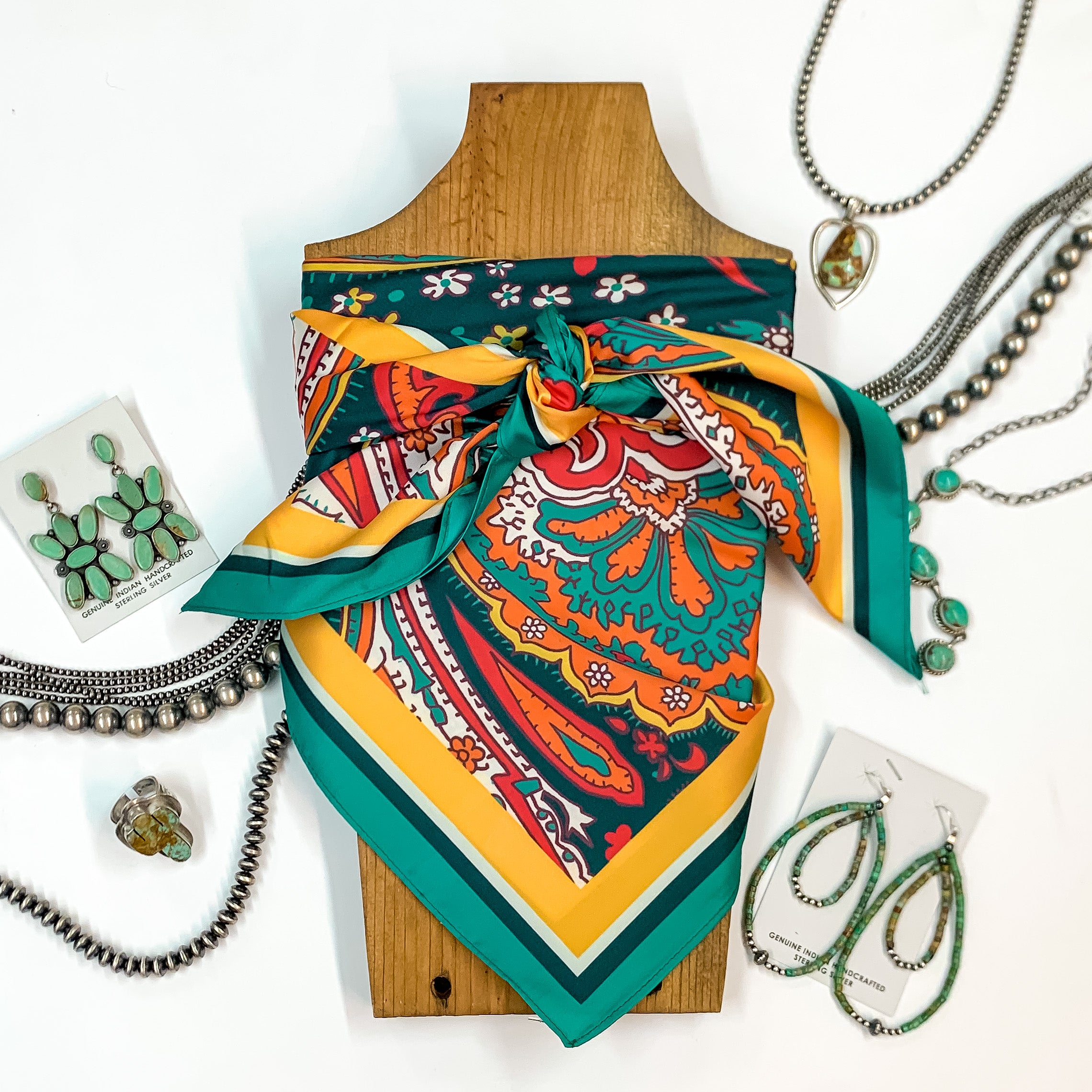 Patterned scarf in Payson Paisley. Scarf is tied around a wooden piece. The scarf and piece of wood is pictured on a white background with Navajo jewelry spread out around it.