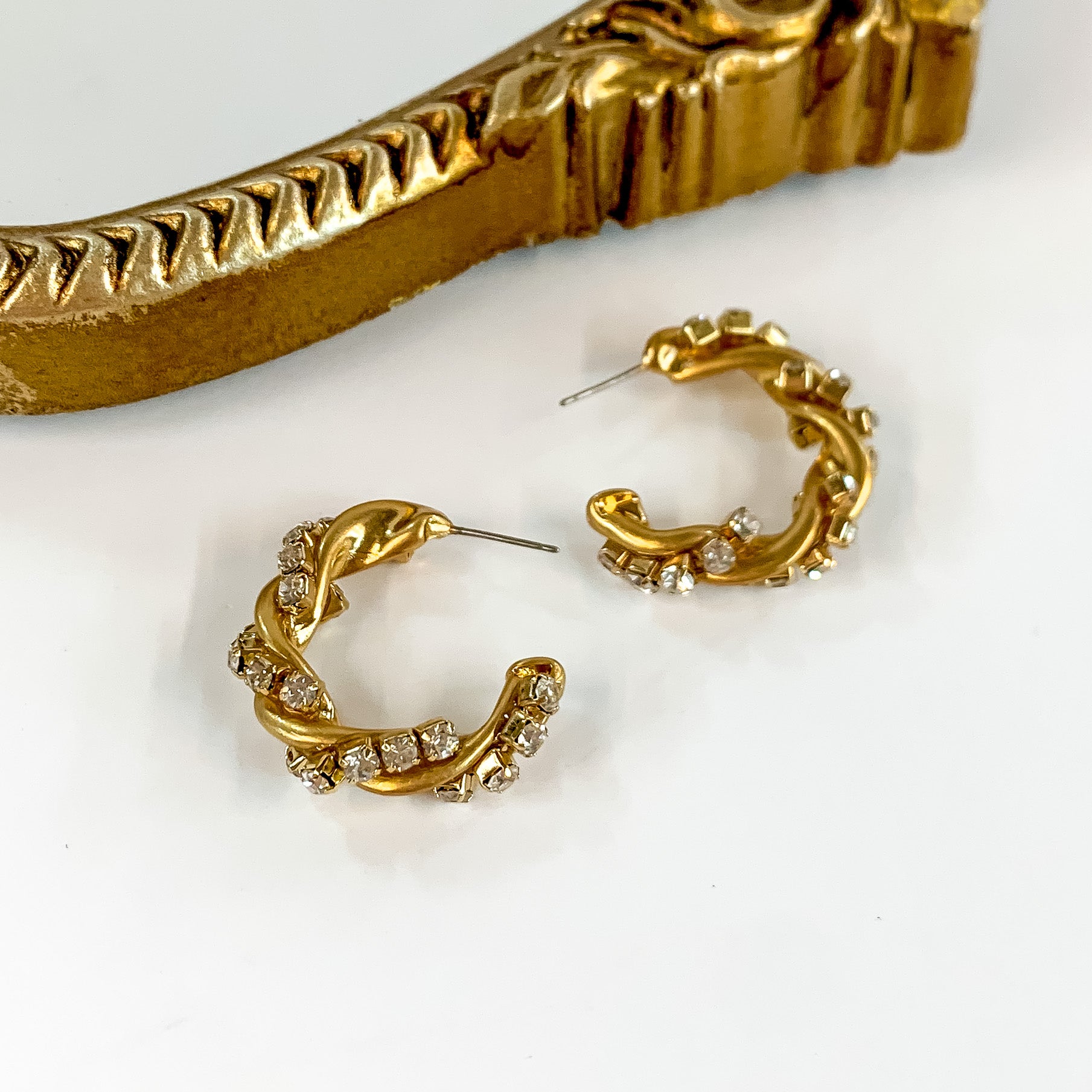 Gold Tone Twisted Hoop Earrings with Clear Crystals. Pictured on a white background with a gold frame behind the earrings.  