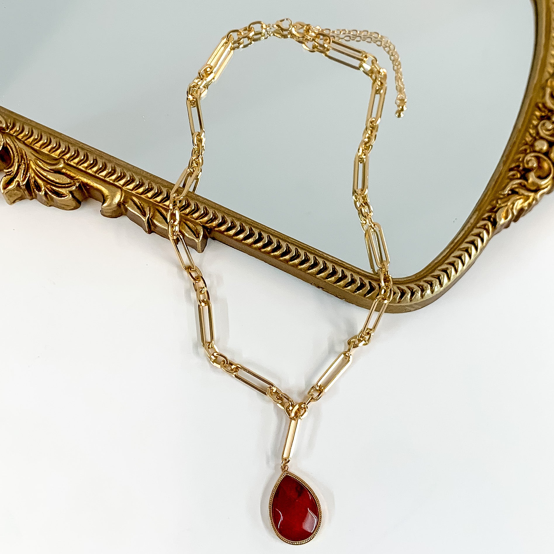 Gold Tone Chain Necklace with Pendant in Maroon. Pictured on a white background with part of the necklace over a mirror.