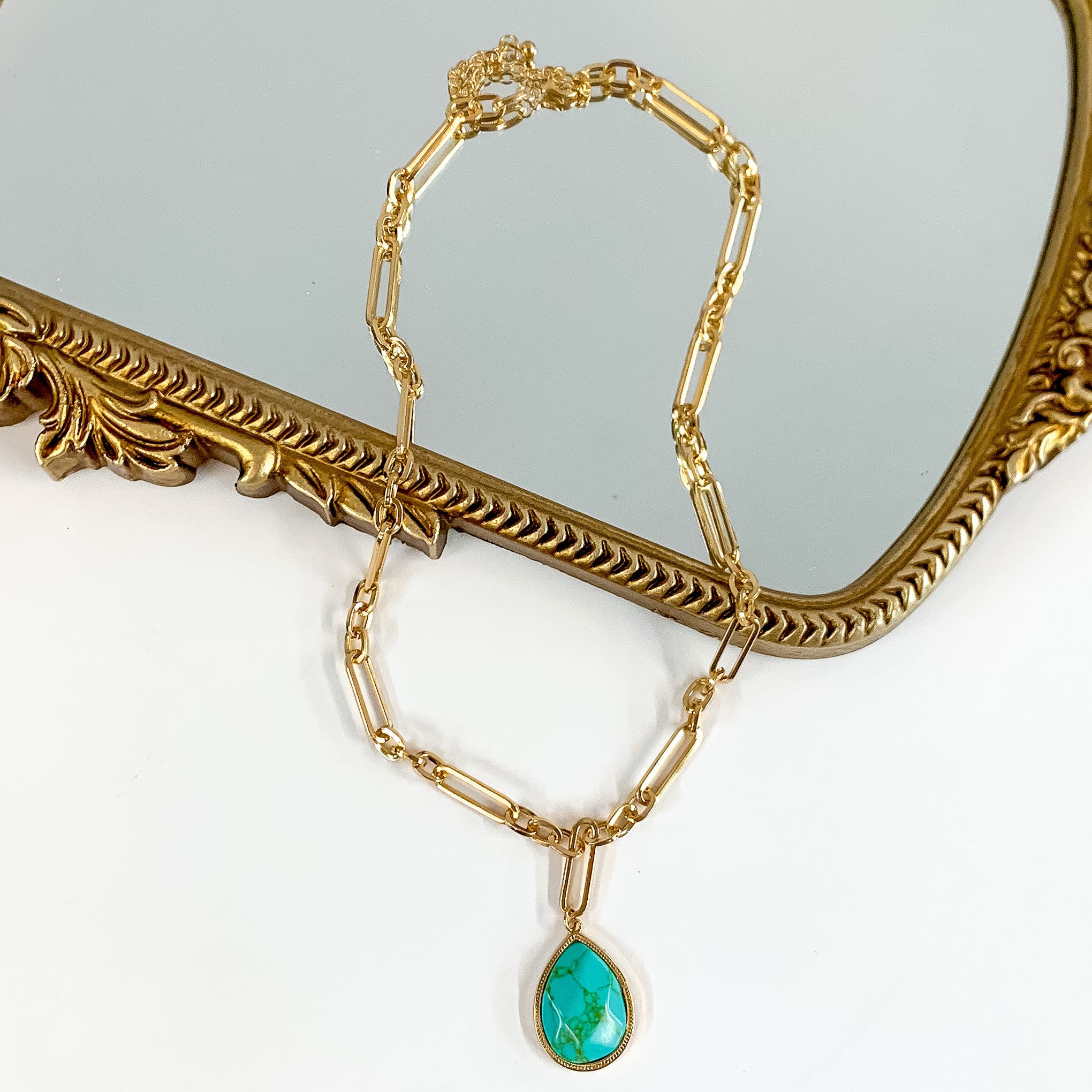 Gold Tone Chain Necklace with Pendant in Turquoise. Pictured on a white background laying halfway on a mirror with a gold trim. 