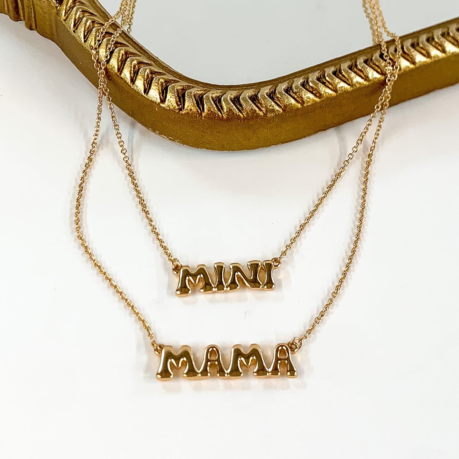 Two gold chain necklaces with word plates. The longer necklace says "MAMA" and the shorter necklace says "MINI." These neckalces are pictured partially laying on a gold mirror on a white background. 