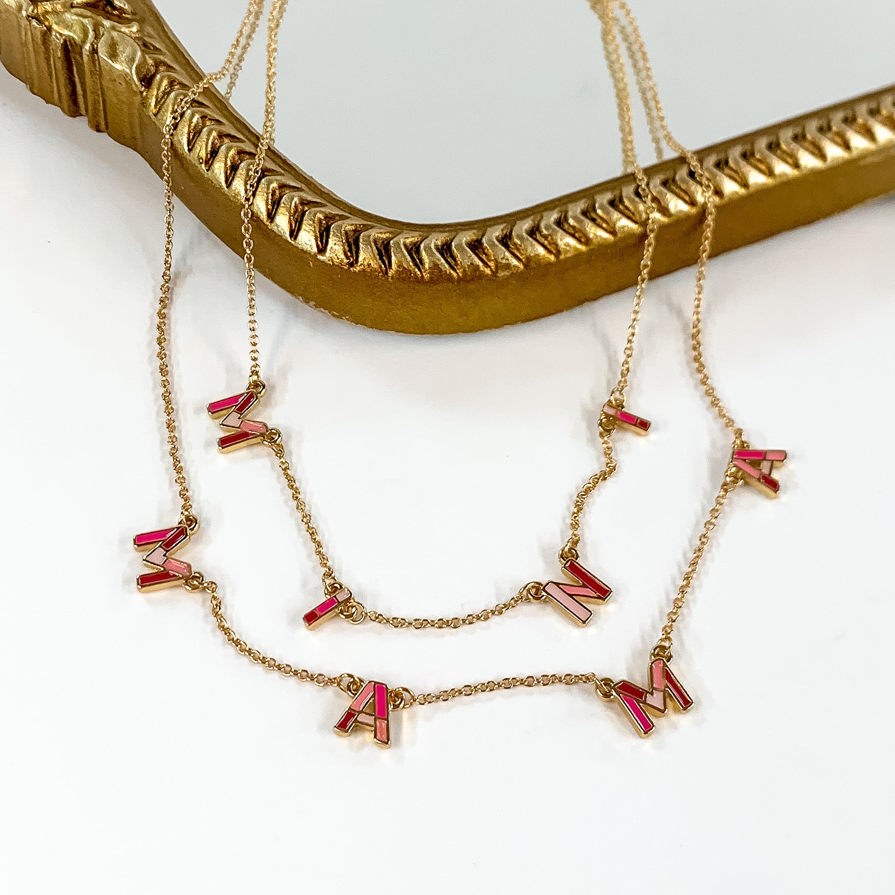 Two gold chain necklaces with pink mix letters. The longer necklace spells out "MAMA" and the shorter necklace says "MINI." These neckalces are pictured partially laying on a gold mirror on a white background. 