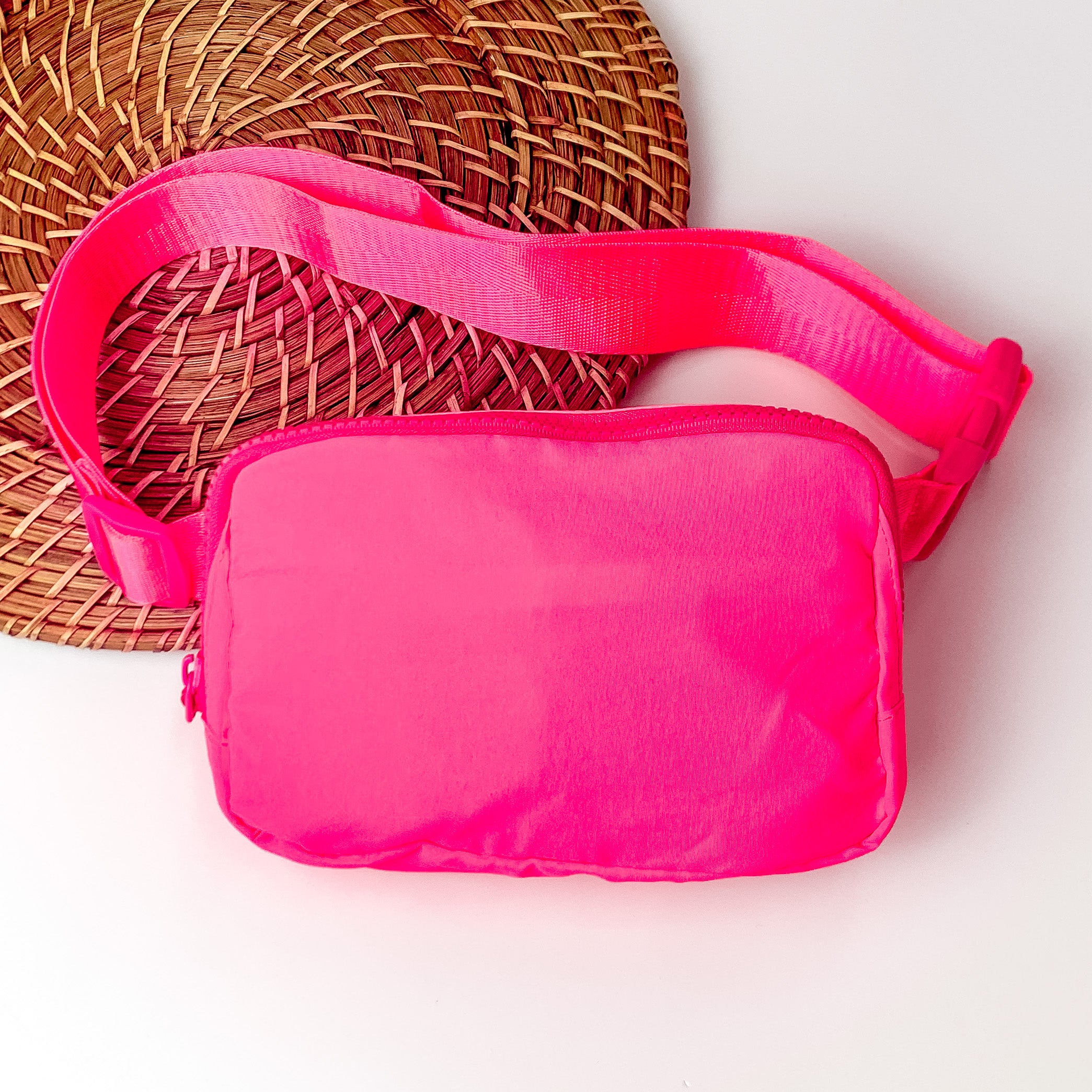 Pictured is a rectangle fanny pack with a top zipper with tassel in pink. This bag also includes a pink strap and pink accents. This bag is pictured on a white and brown patterned background.