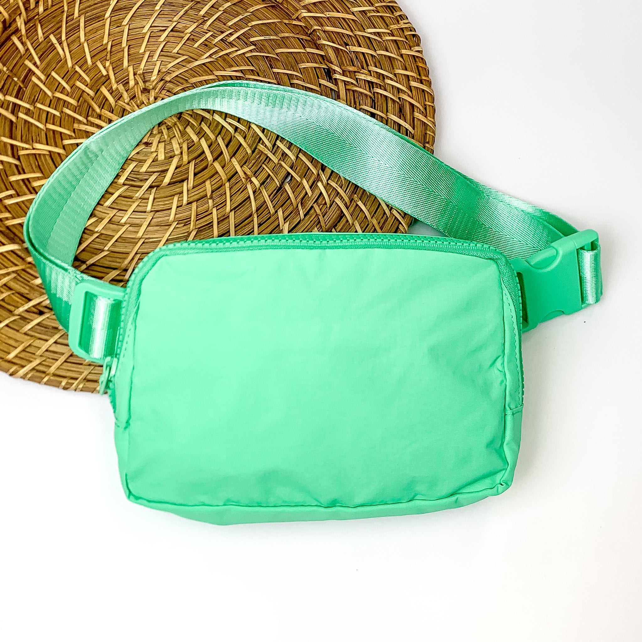 Pictured is a rectangle fanny pack with a top zipper with tassel in turquoise. This bag also includes a turquoise strap and turquoise accents. This bag is pictured on a white and brown patterned background.