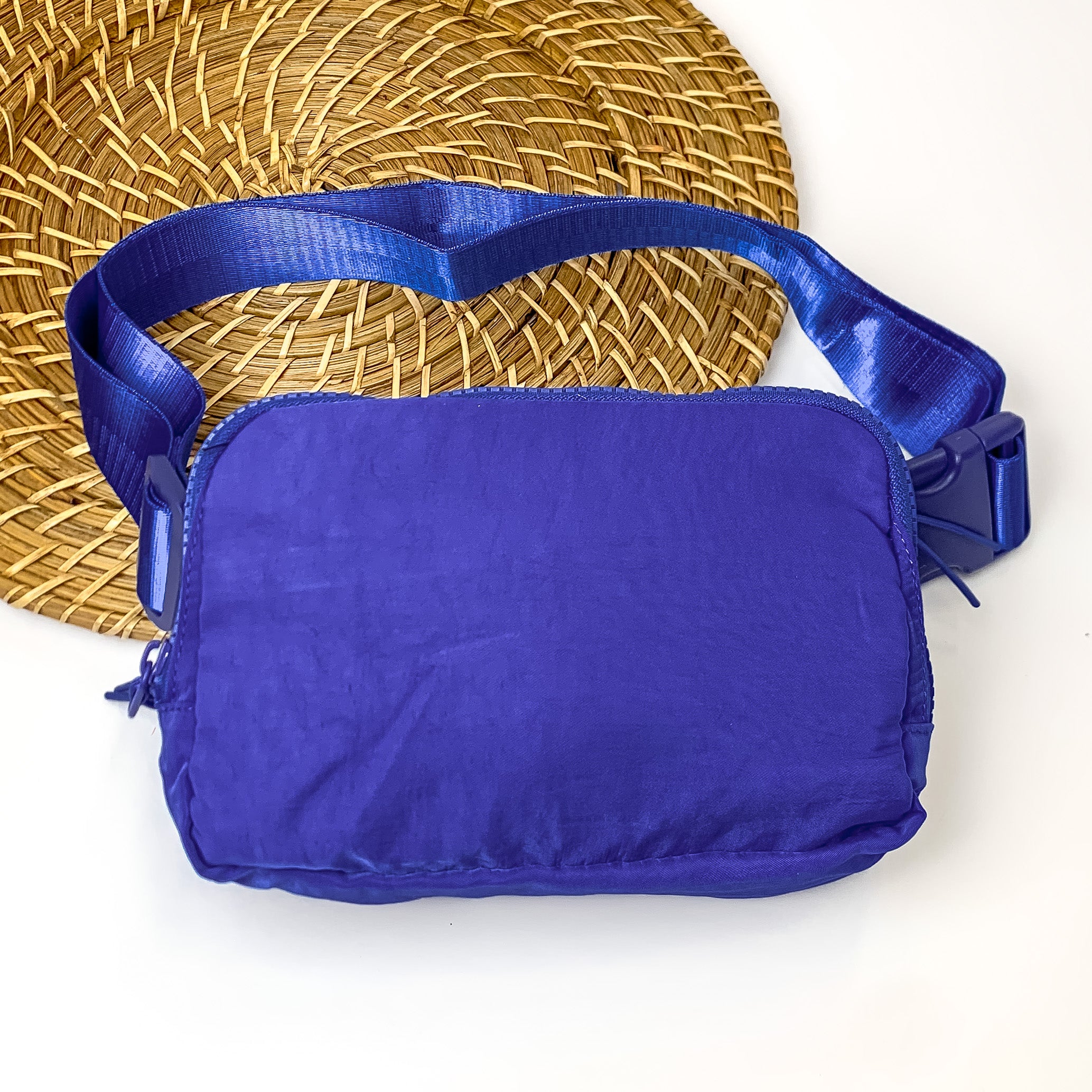Pictured is a rectangle fanny pack with a top zipper with tassel in royal blue. This bag also includes a royal blue strap and royal blue accents. This bag is pictured on a white and brown patterned background.