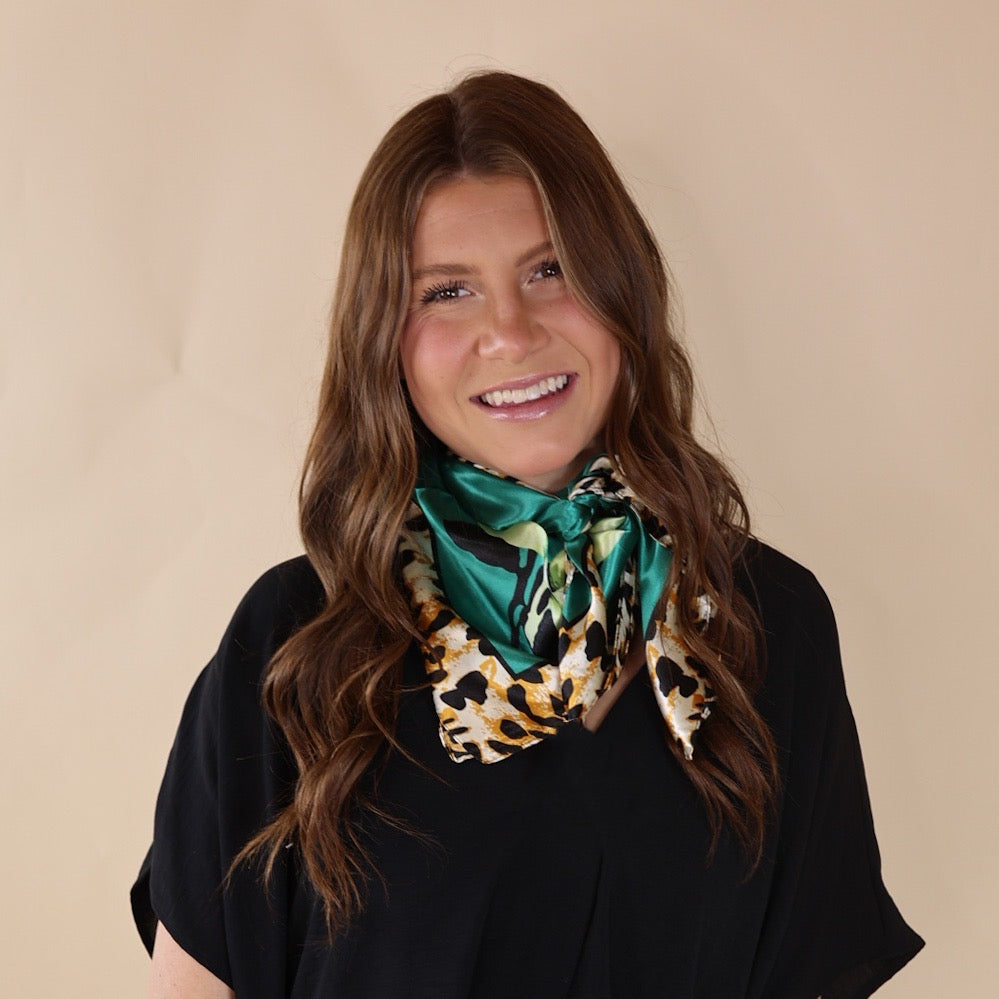 Brunette model is wearing a white drop shoulder top with a emerald green scarf with a cheetah print border wrapped and tied around her neck. She is pictured in front of a beige background.