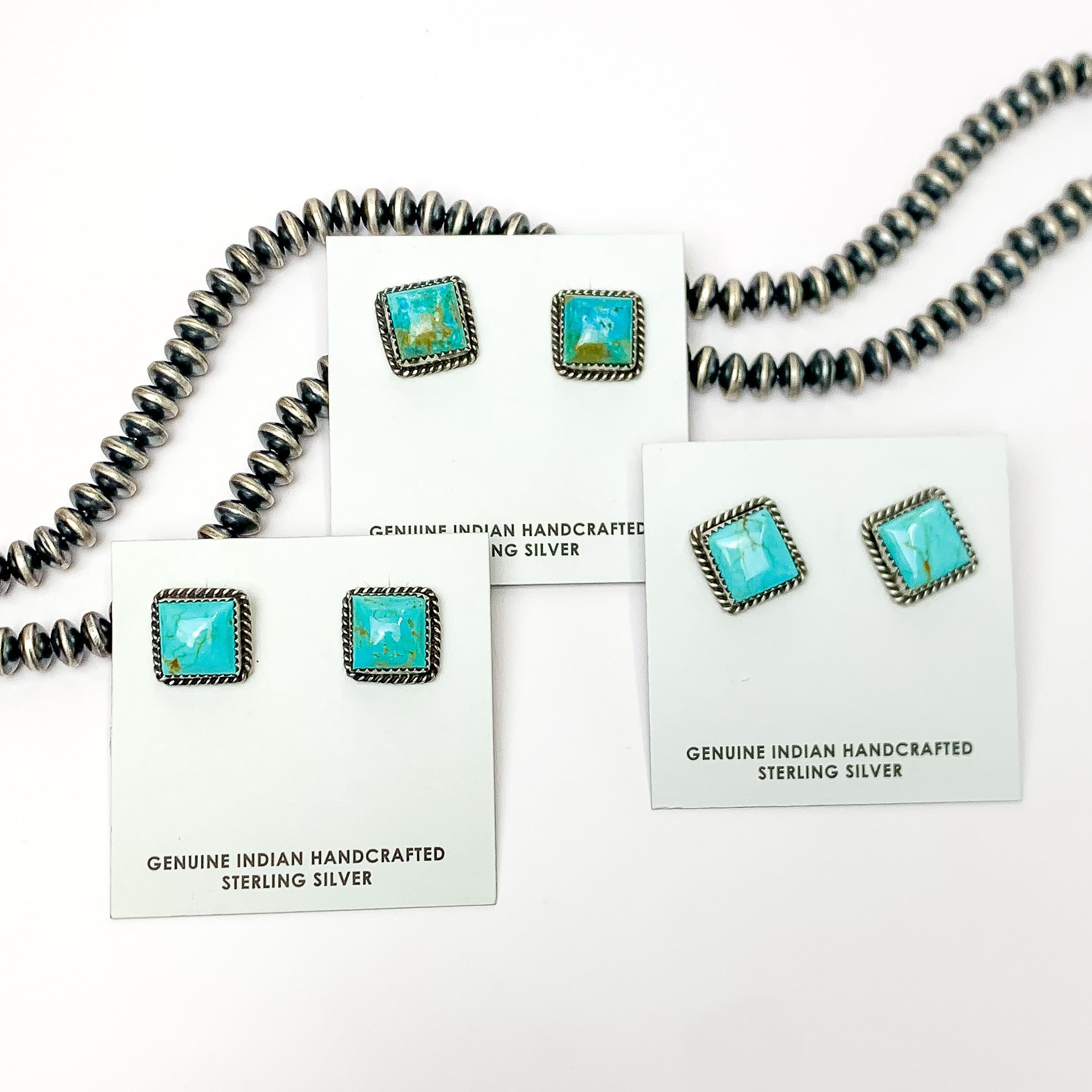 Pictured are three pairs of square, turquoise stud earrings on white earring holders. These earrings are pictured in front of silver beads on a white background. 