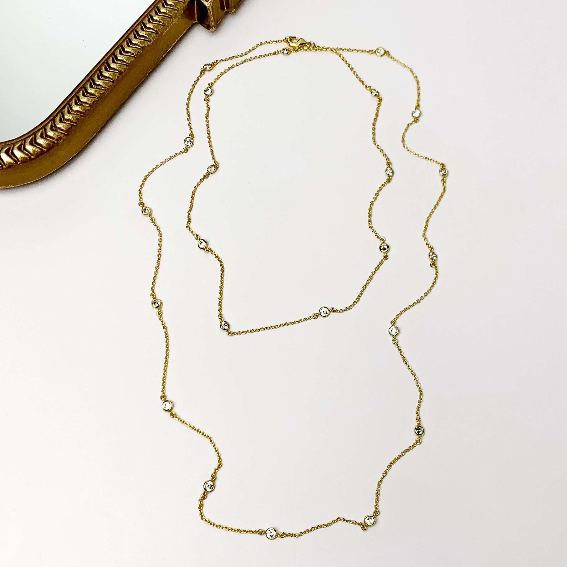 Simple Long Gold Tone Necklace With Clear Crystals. Pictured on a white background with a wood piece behind the necklace.