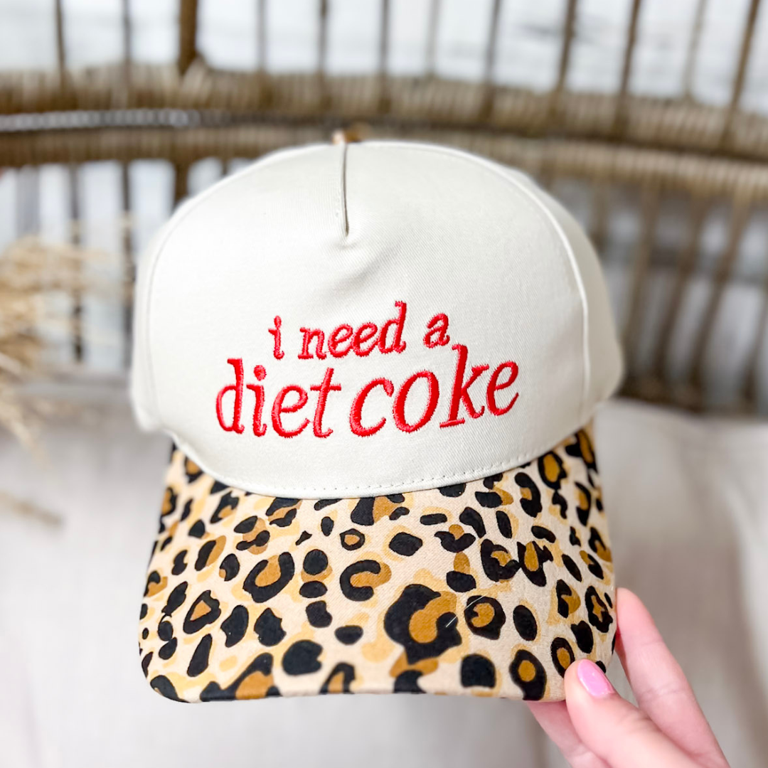 I Need a Diet Coke embroidered two-tone canvas trucker cap featuring a cream crown, bold red lettering, leopard-print brim, and adjustable snapback closure.