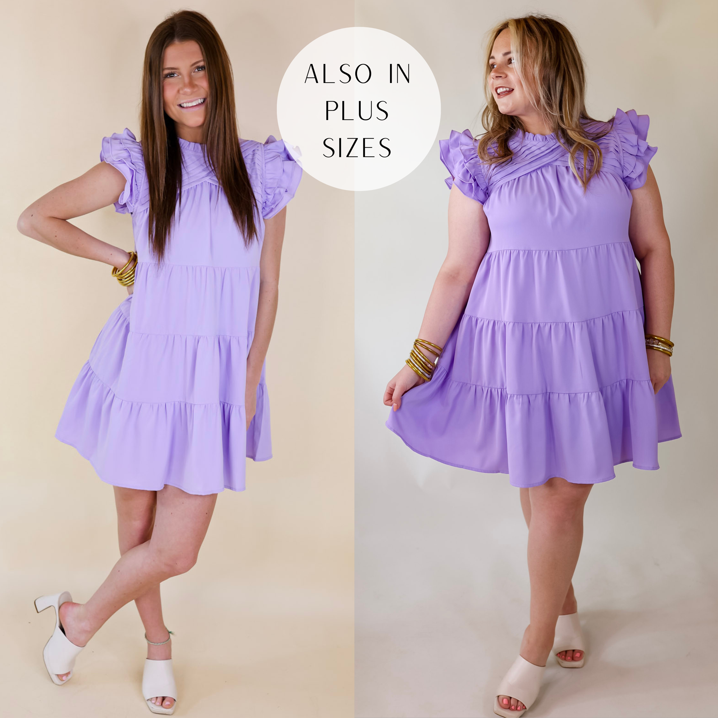 Model is wearing a lavender purple dress with pleated detailing and ruffle cap sleeves. Model has this paired with white heels and gold jewelry.