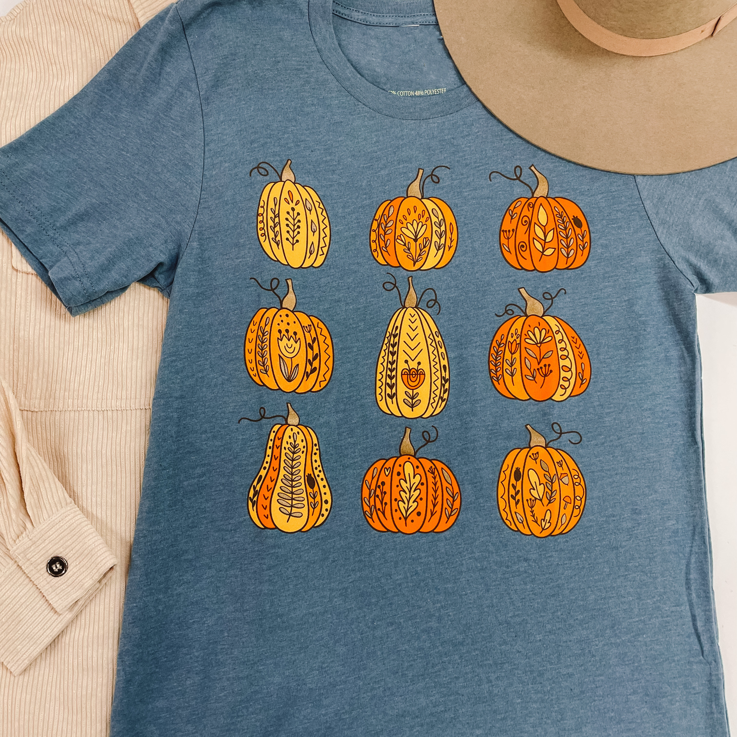 A short sleeve graphic tee with three rows of three pumpkins that have a flower on the front. Pictured on a white background with a beige shacket and a mushroom felt hat.