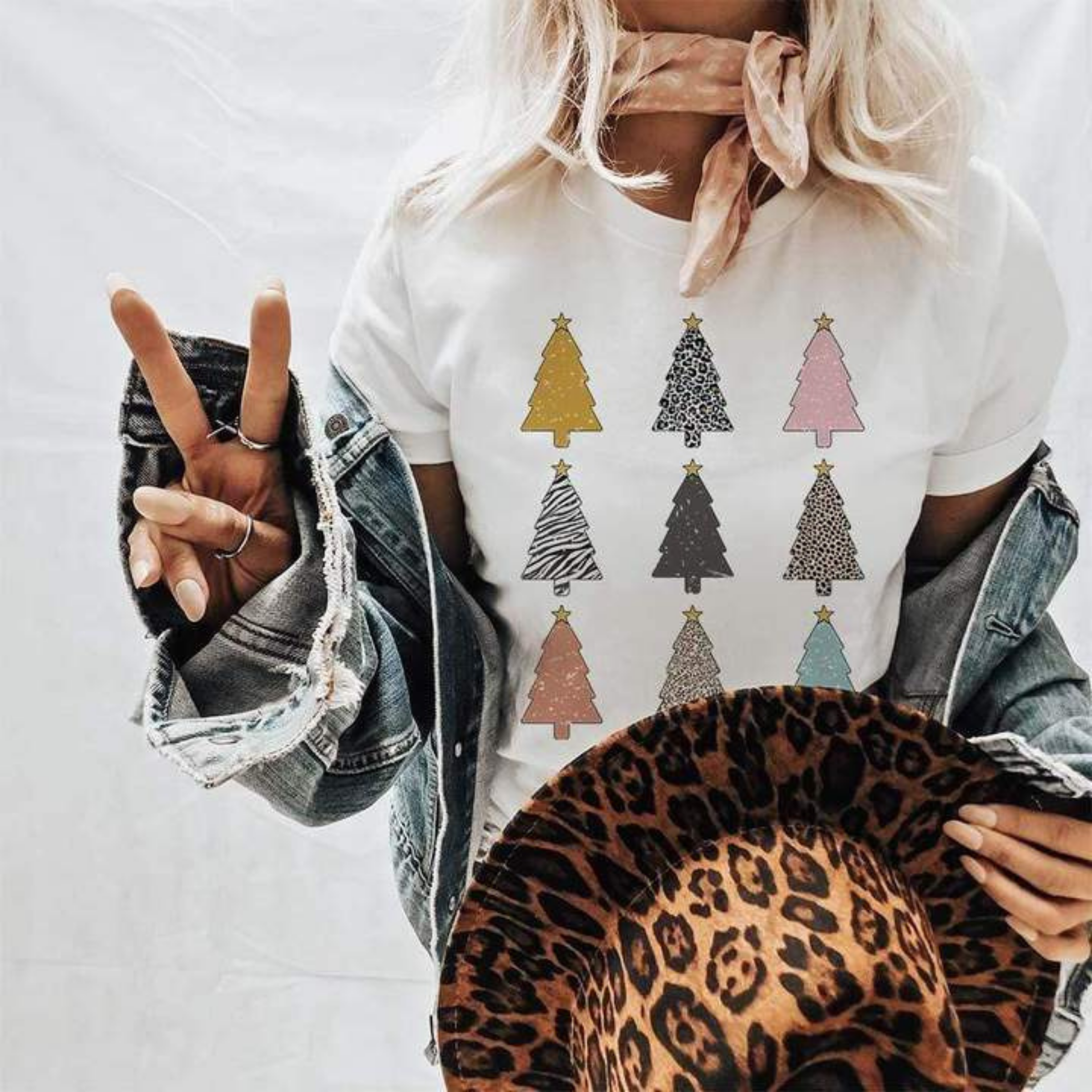 Model is wearing a white short sleeve tee with 3 rows of 3 different print Christmas trees. Model has it paired with a denim jacket, leopard hat, and wild rag tied around the neck.