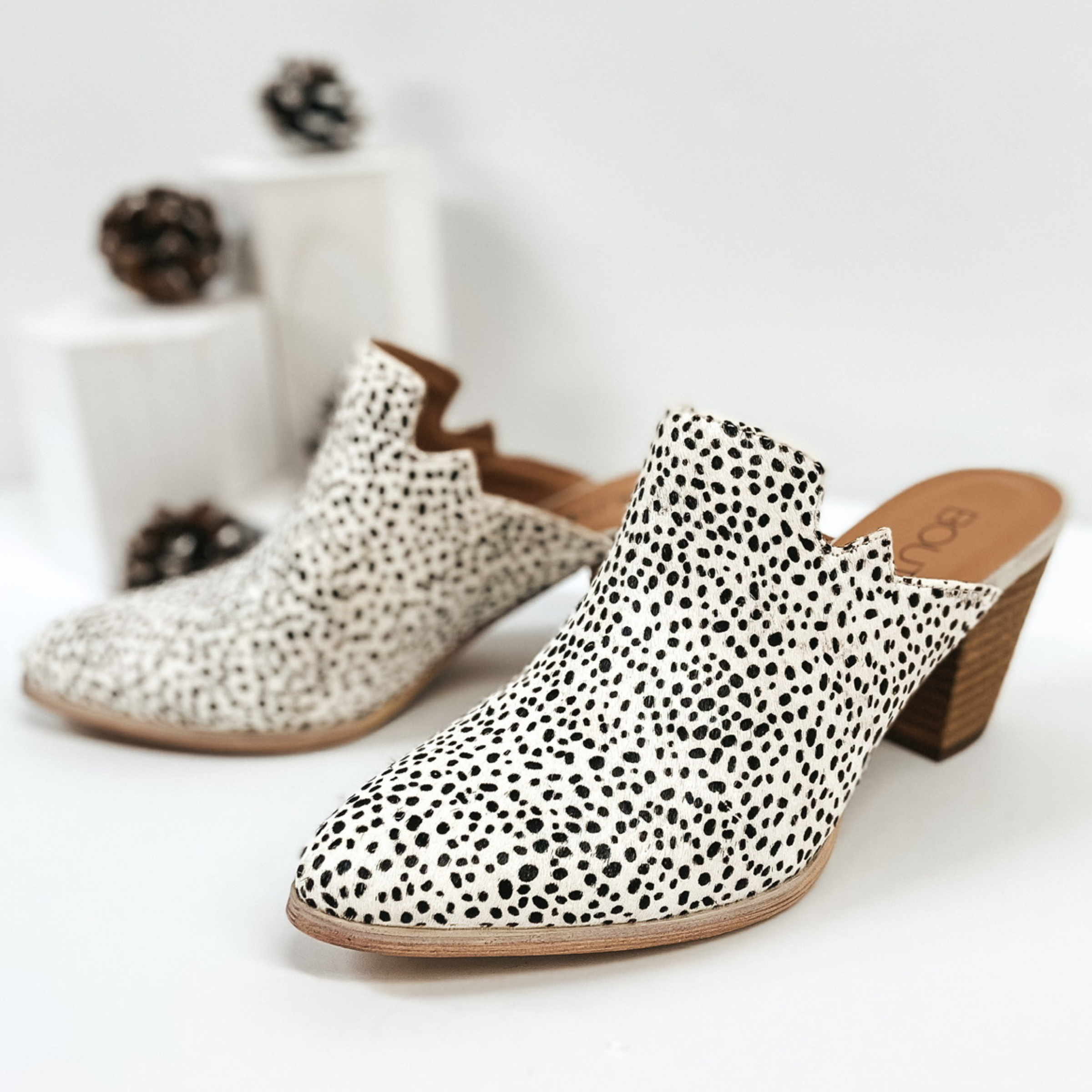 White speckled with black hair on hide heeled mules. Pictured on white background with pinecones.