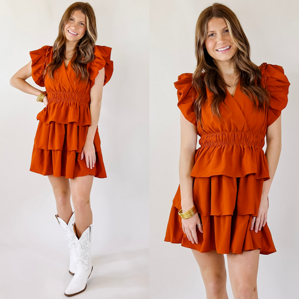 Model is wearing a short orange dress featuring ruffled sleeves, cross front, cinched waist, a tiered skirt.