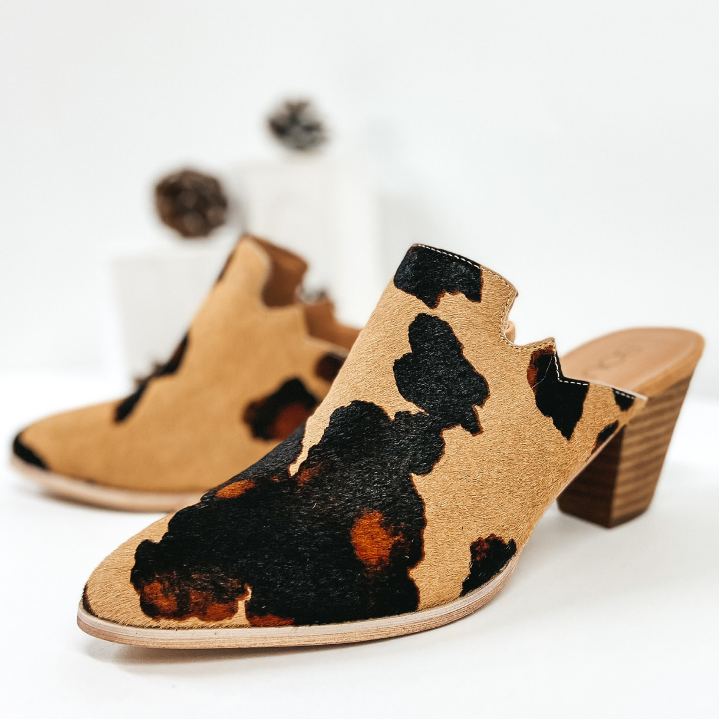 Tan cow print hair on hide heeled mules. Pictured on white background with pinecones.