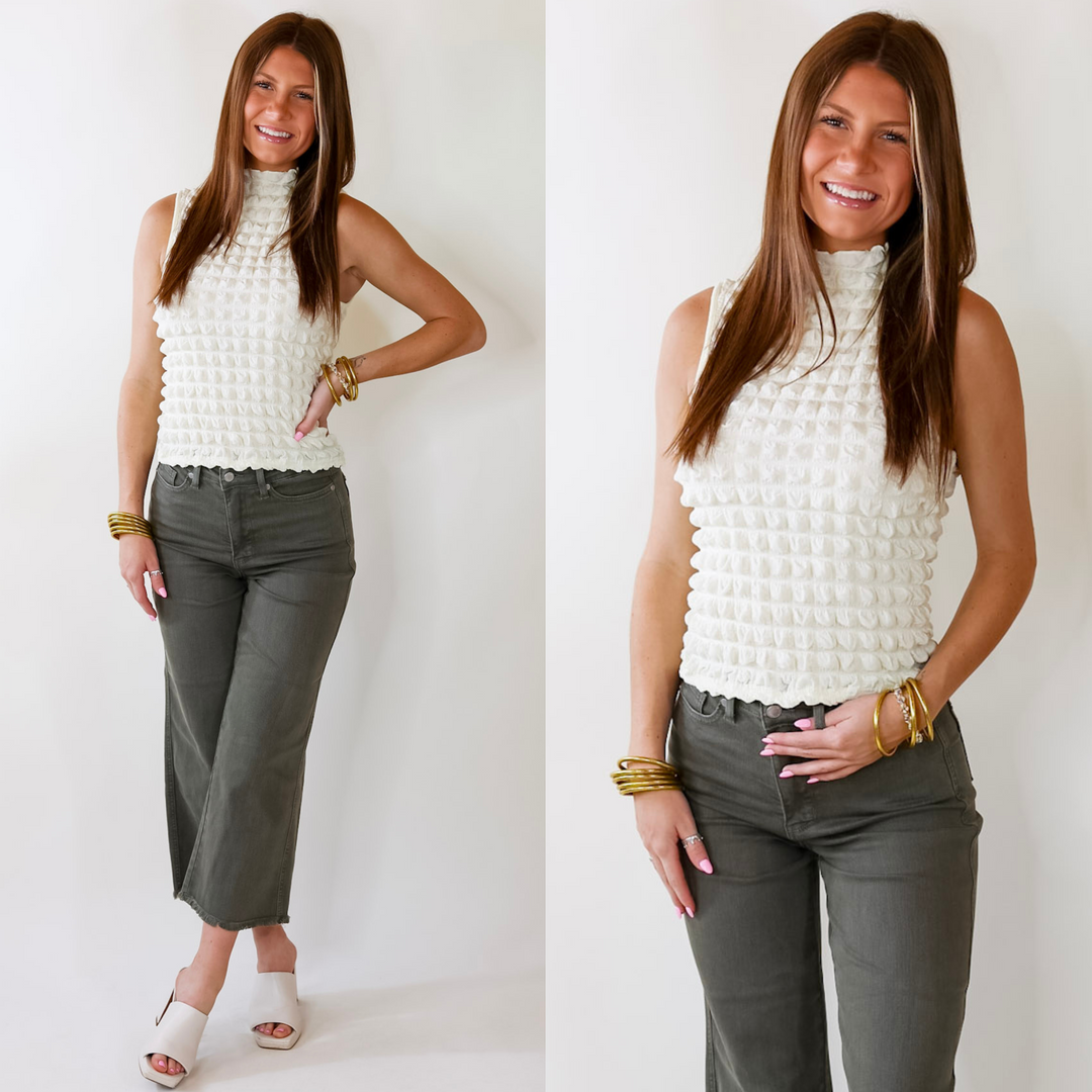 Model is wearing an ivory mock neck tank top. Model has it paired with olive green jeans, ivory heels, and gold jewelry.