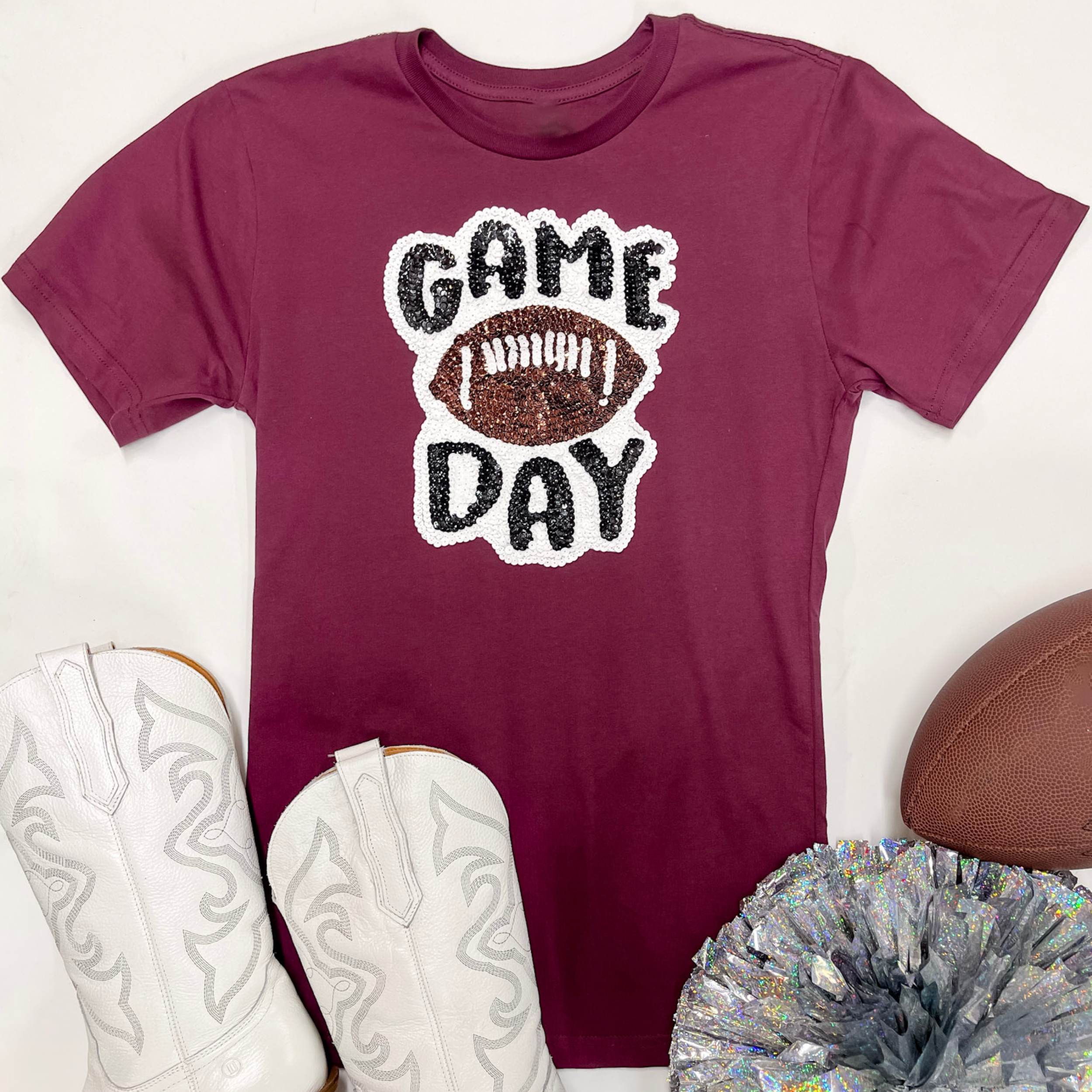 A maroon short sleeve graphic tee with a sequin patch. pictured on a white background with a silver pom pom and white boots.