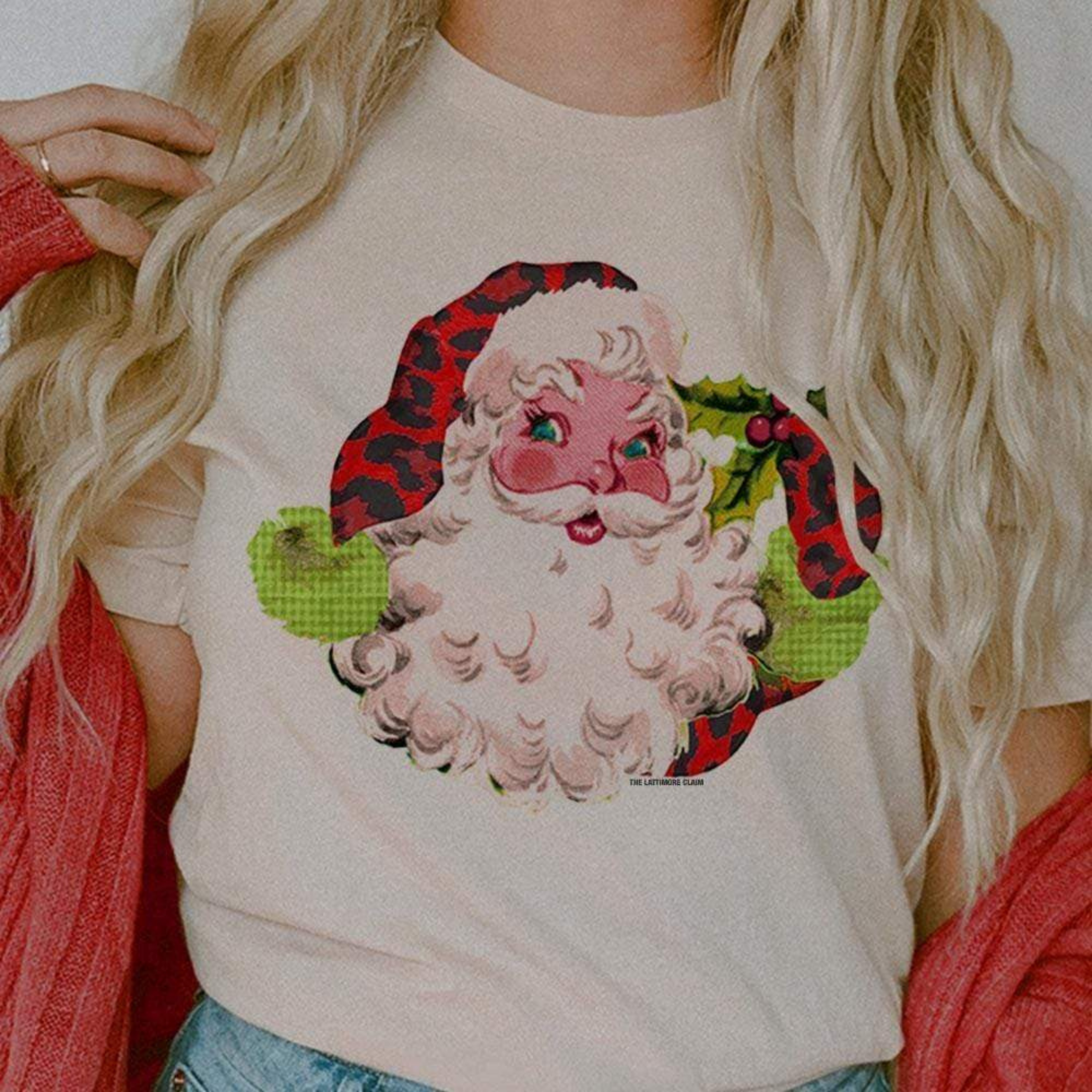 This cream tee includes a crew neckline, short sleeves, and a vintage Santa Clause graphic. Santa is wearing a red leopard print and green hat and glove. The model has this shown with rolled sleeves and a red cardigan. 