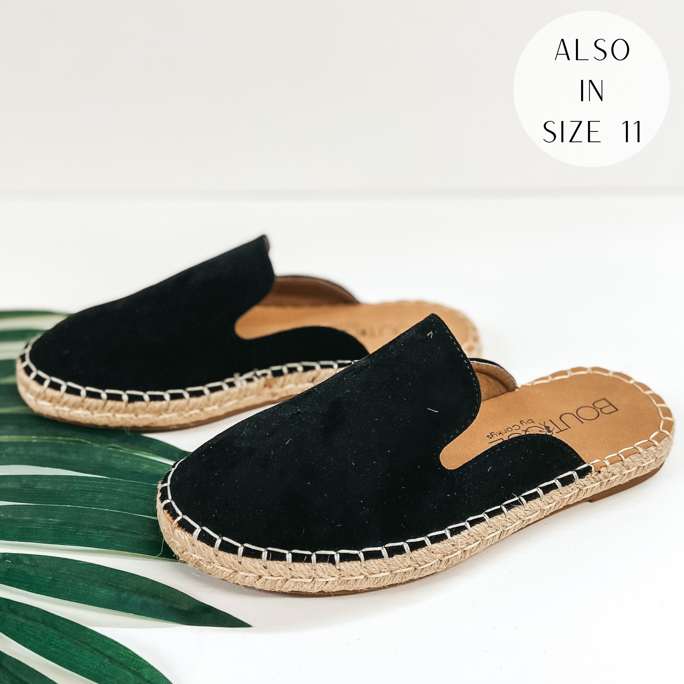 A pair of slip on espadrille shoes that have a black upper. Pictured on white background with a green palm leaf.