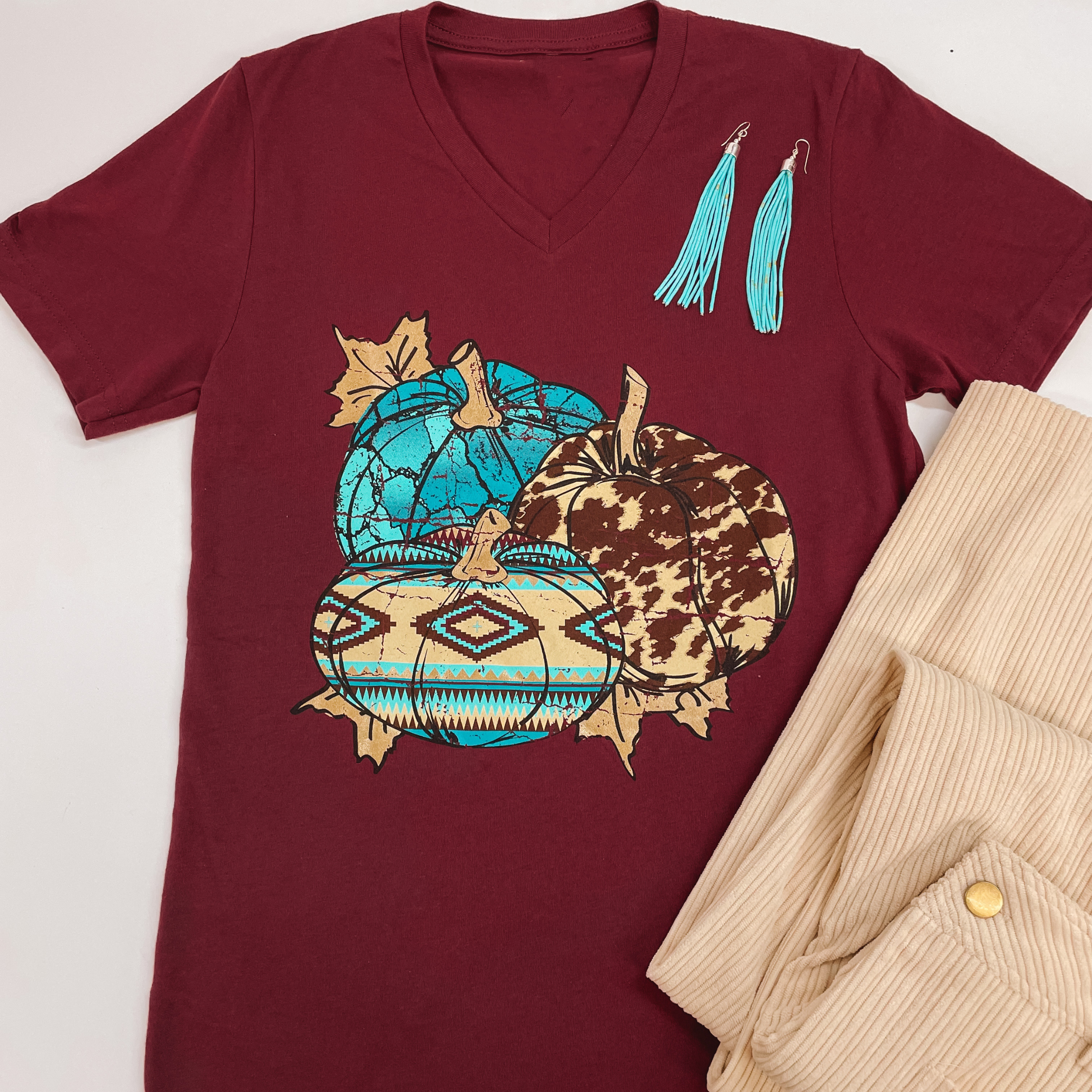 A short sleeve graphic tee with a v neckline and a graphic of a cow print pumpkin, turquoise pumpkin, and Aztec print pumpkin. Pictured on a white background with beige corduroy pants and turquoise earrings.