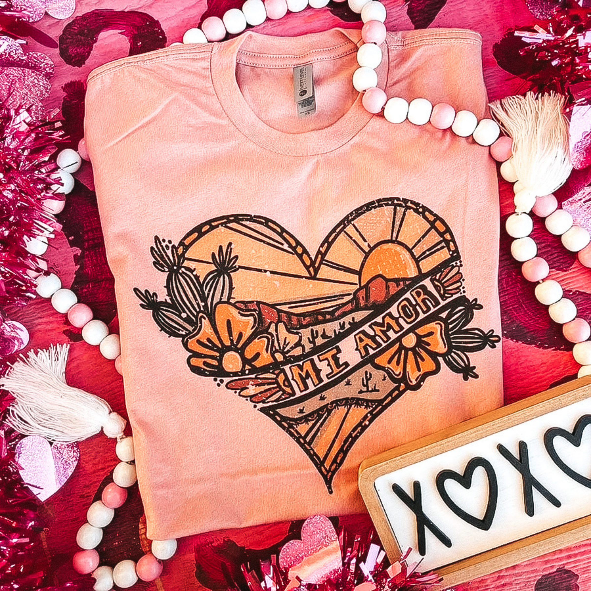 A light coral tee shirt with a desert heart graphic. Pictured on a pink background with beads, hearts, and tinsel.