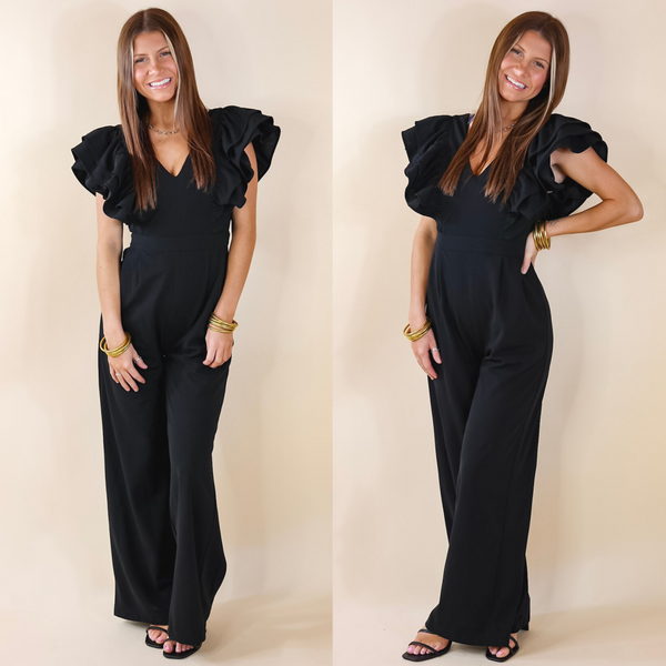 Model is wearing a black jumpsuit with ruffle sleeves and a v neckline. Model has it paired with black heels and gold jewelry.