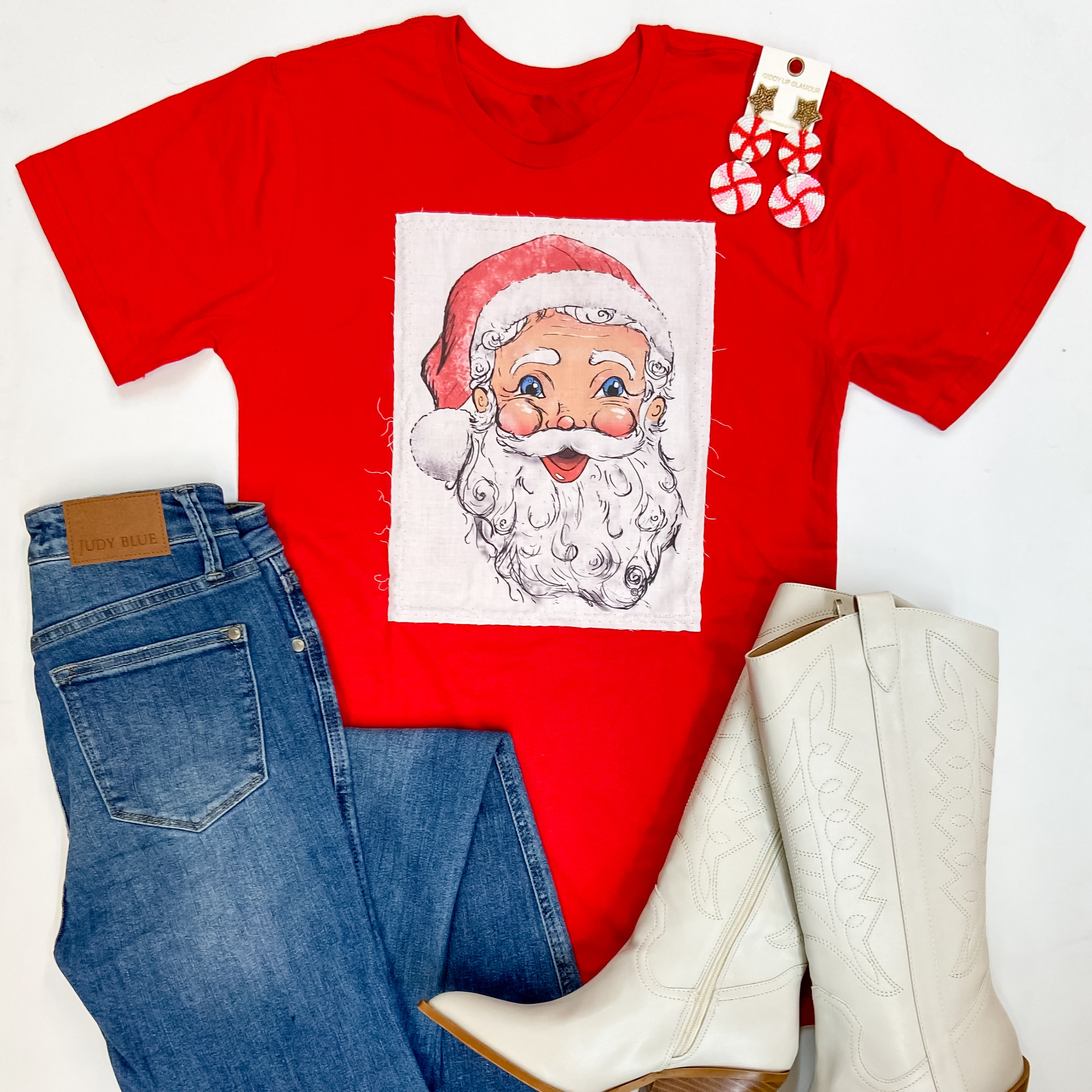 A red short sleeve tee shirt with a patch of Santa Clause. Pictured on a white background with peppermint earrings, flare jeans, and ivory booties.