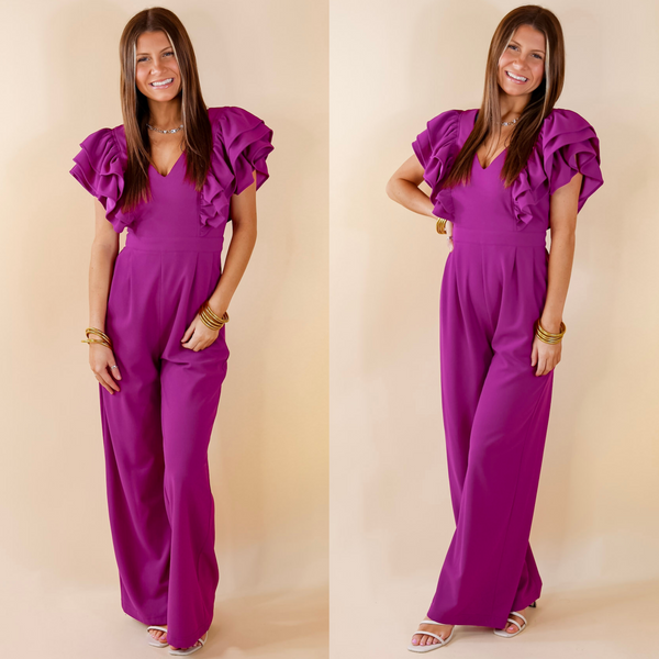Model is wearing a purple jumpsuit with ruffle detail sleeves and a v neckline. Model has it paired with white heels and gold jewelry.