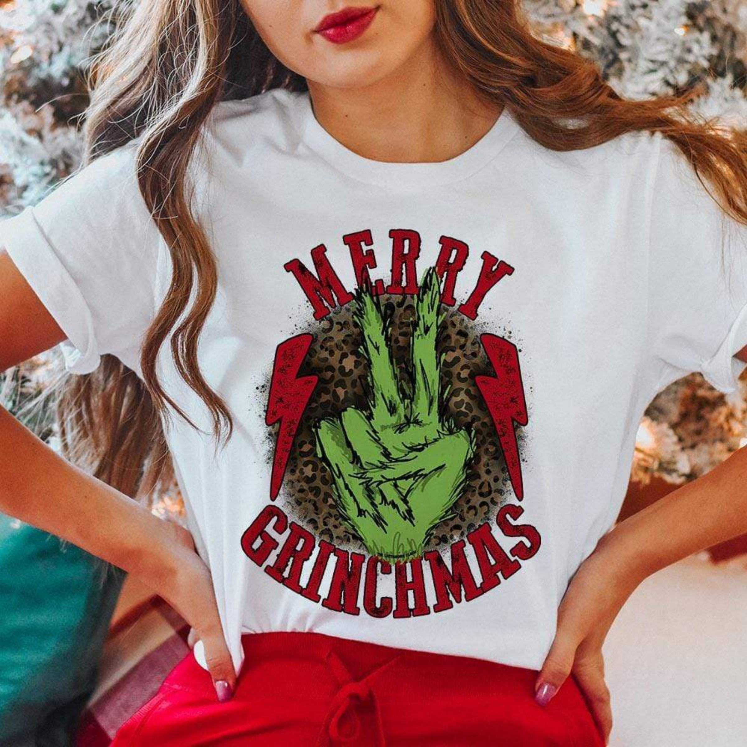 This white tee includes a crew neckline, short sleeves, and a cute Christmas graphic that says "Merry Grinchmas" in red font, a Grinch hand holding up the peace sign, and two red lightning bolts on the outside of the Grinch hand. This graphic also has a leopard print behind it. The model has this graphic tee styled with rolled sleeves and red pants. 