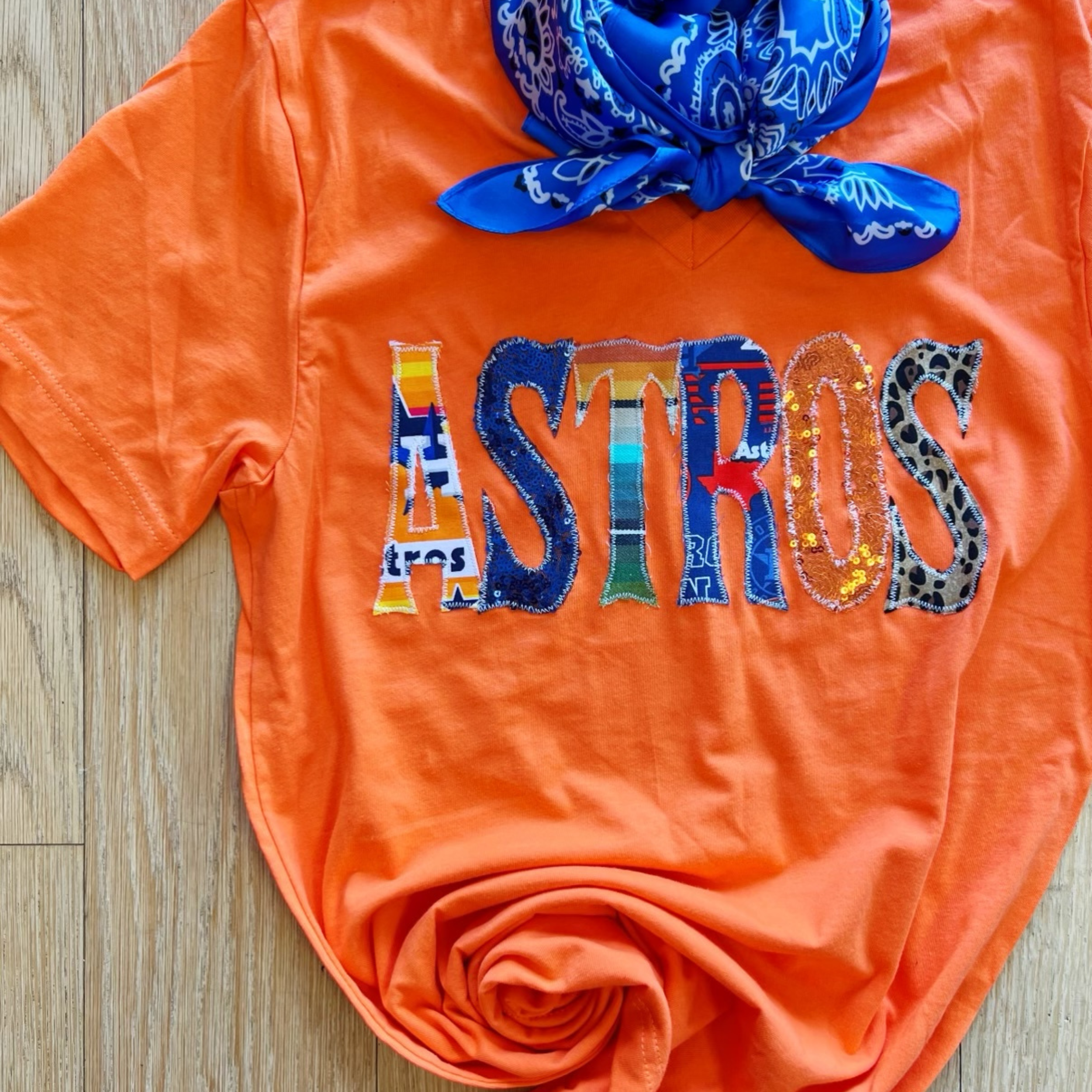 An orange graphic tee with short sleeves, a v neckline, and mix print patch letters that spell "ASTROS." This tee is pictured on a wooden background with a blue bandana. 