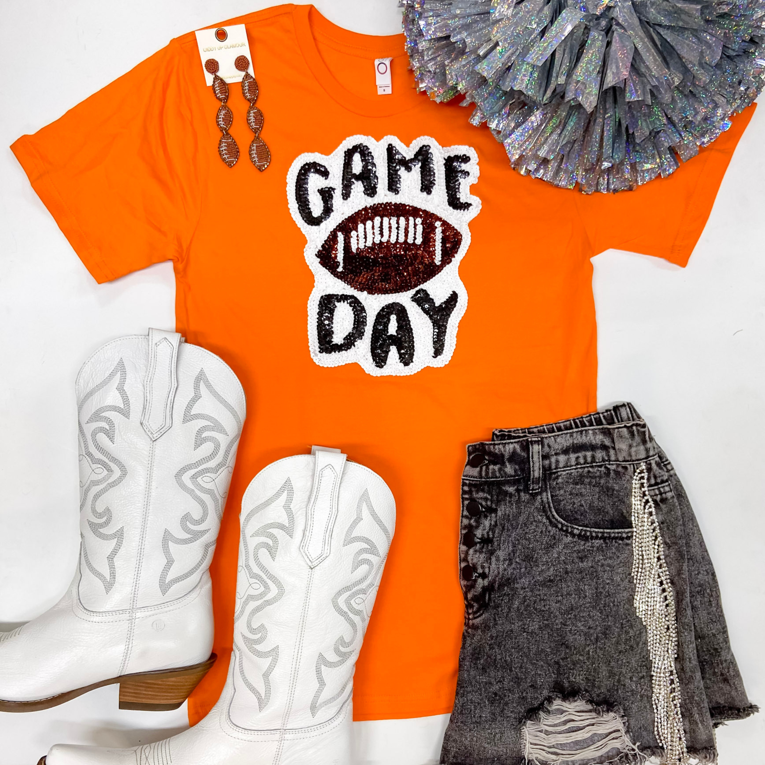 An orange short sleeve tee shirt with a gameday sequin patch on the front. It is laid on a white background with white boots, black shorts, and football earrings.