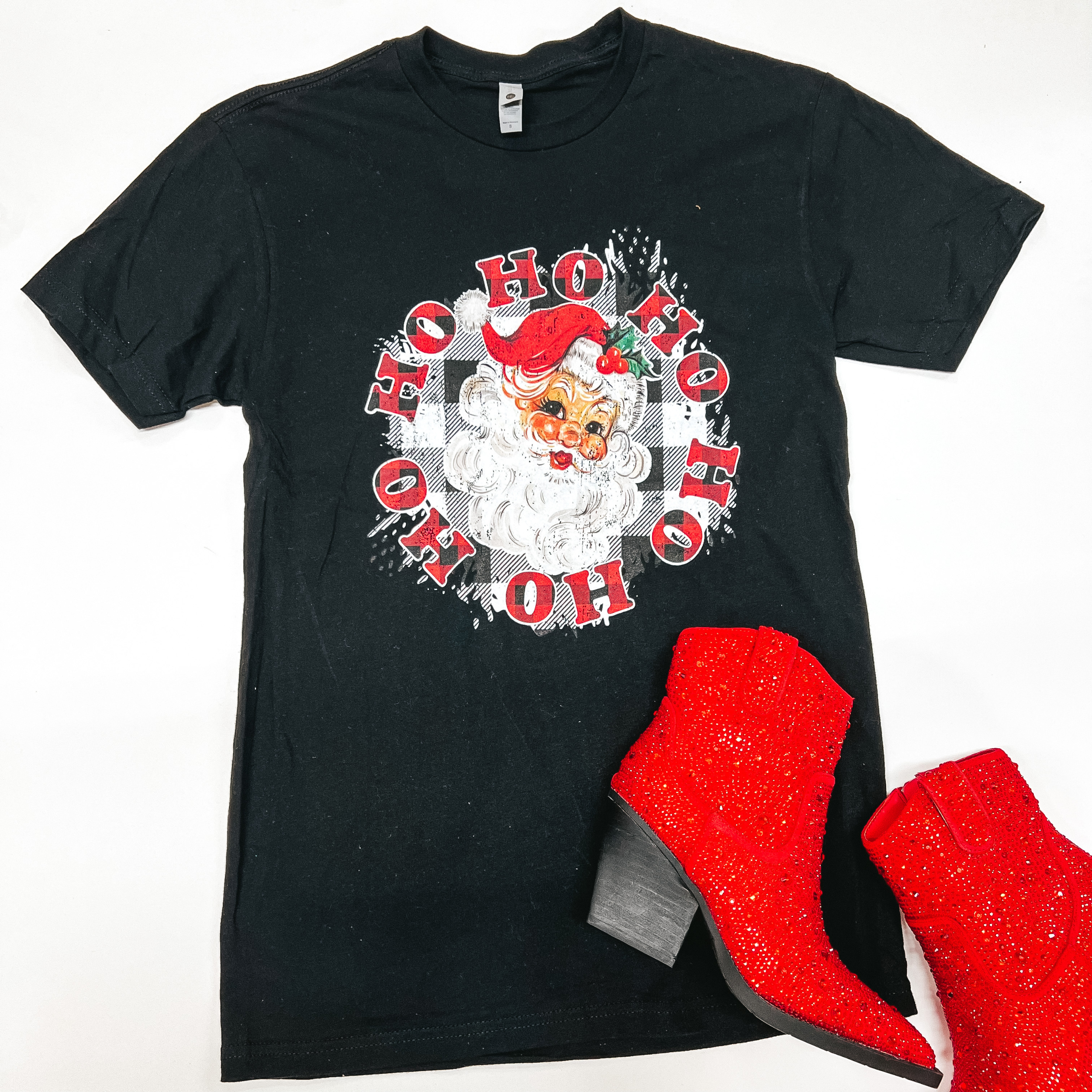 This black tee includes short sleeves, a crew neckline, and a fun Christmas graphic of a classic Santa Clause on a black and white gingham background with the words "HO HO HO HO HO HO", in an uppercase font, in a circle around him in red and black gingham. This tee is shown as a flatlay on a white background and it is paired with red, sparkly booties. 