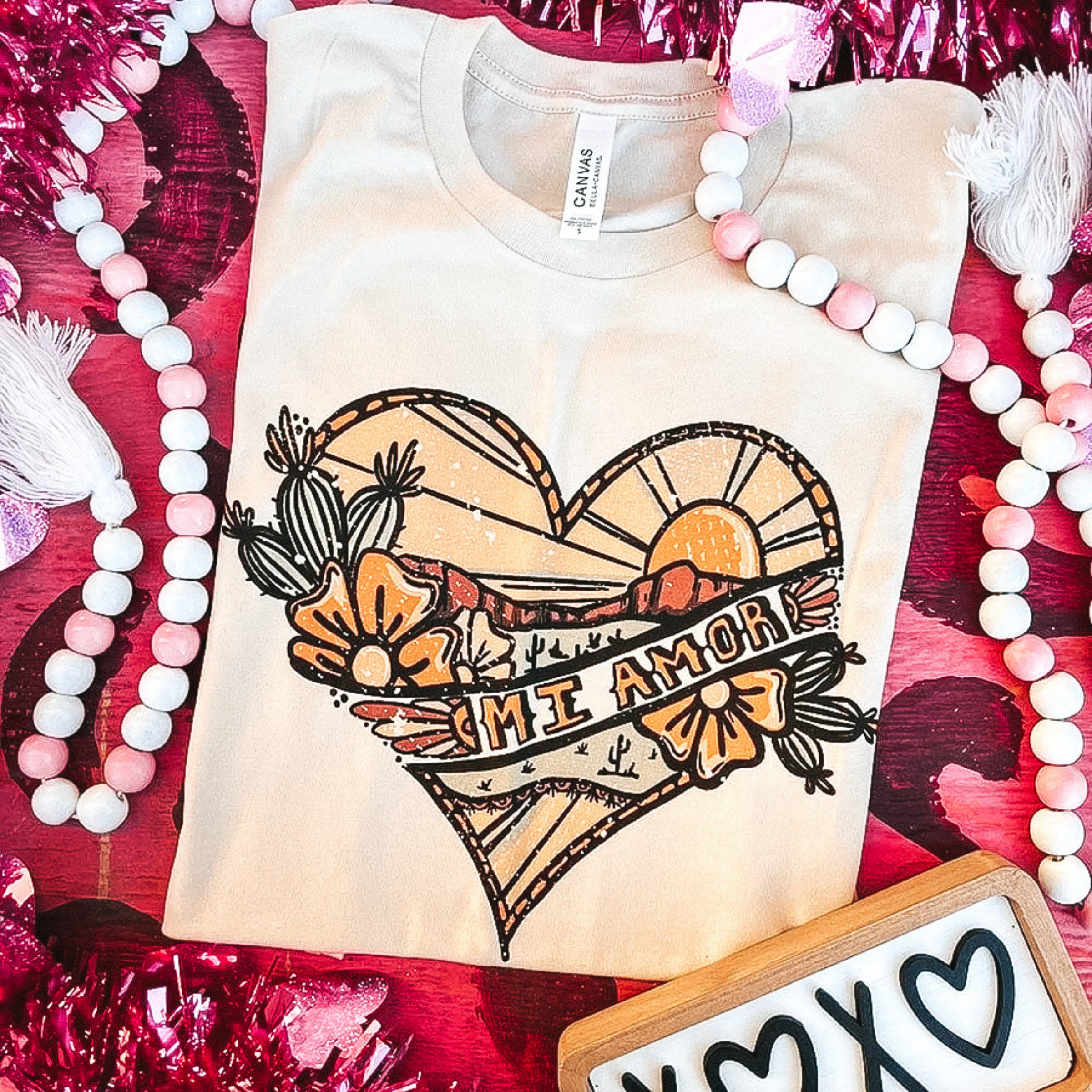 An ivory tee shirt with a desert heart graphic. Pictured on a pink background with beads, hearts, and tinsel.