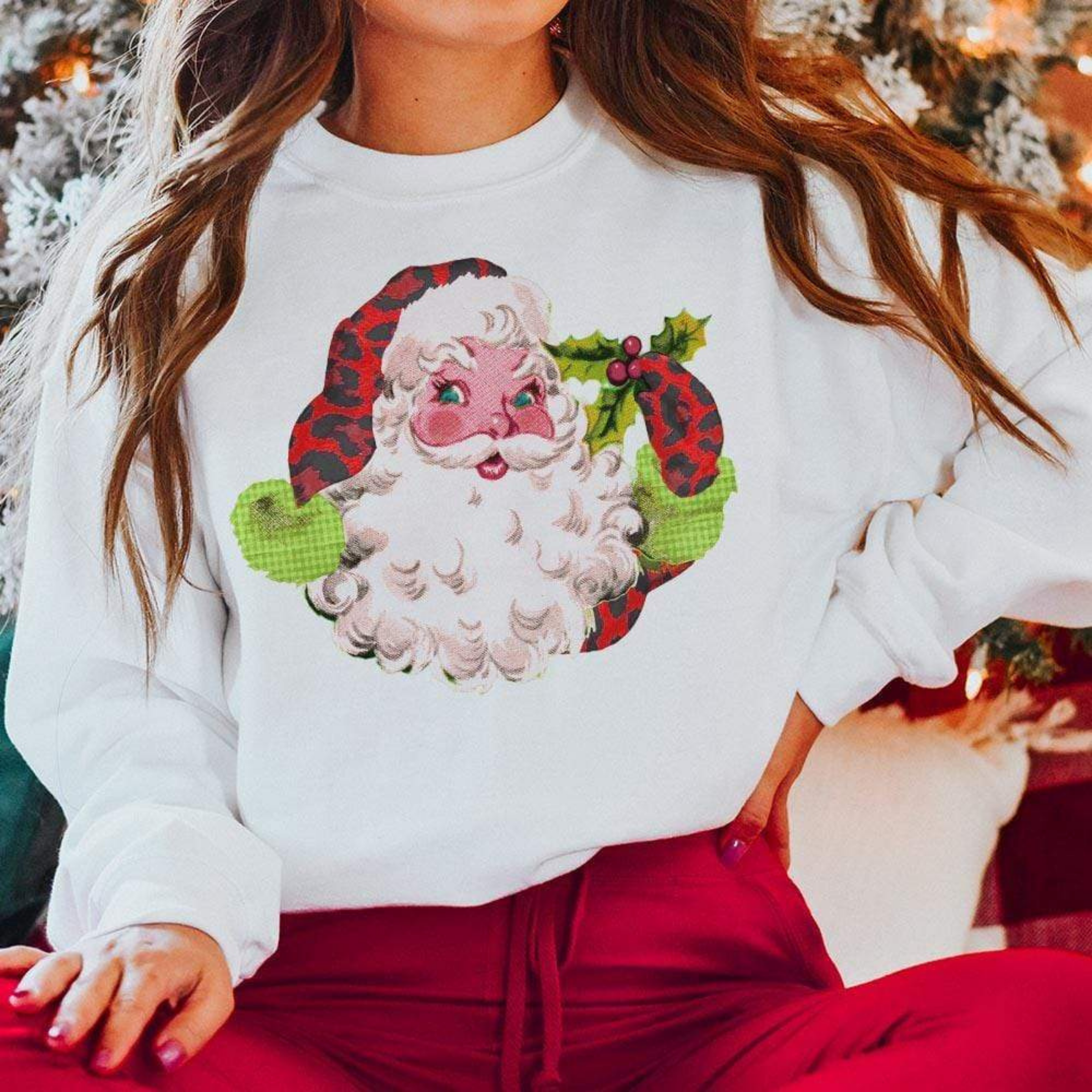 Model is sitting criss-cross with a white sweatshirt on. This sweatshirt has a picture of Santa Clause holding a mistletoe and wearing a red leopard hat and glove. She has it paired with red pants. 
