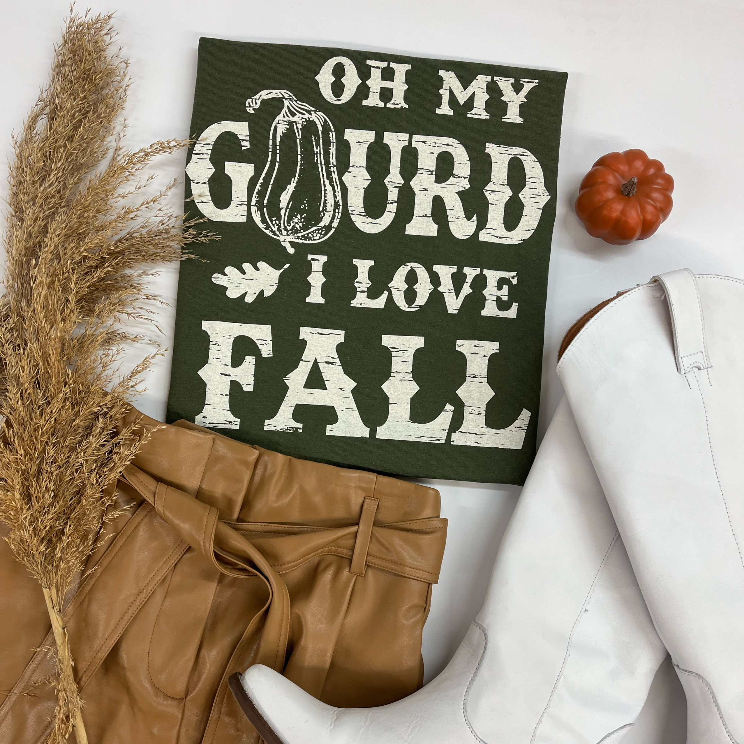A folded tee shirt that is moss green. The t-shirt has white lettering that says  "Oh my Gourd I love fall".  Pictured on a white background with tan faux leather shorts and white boots. 