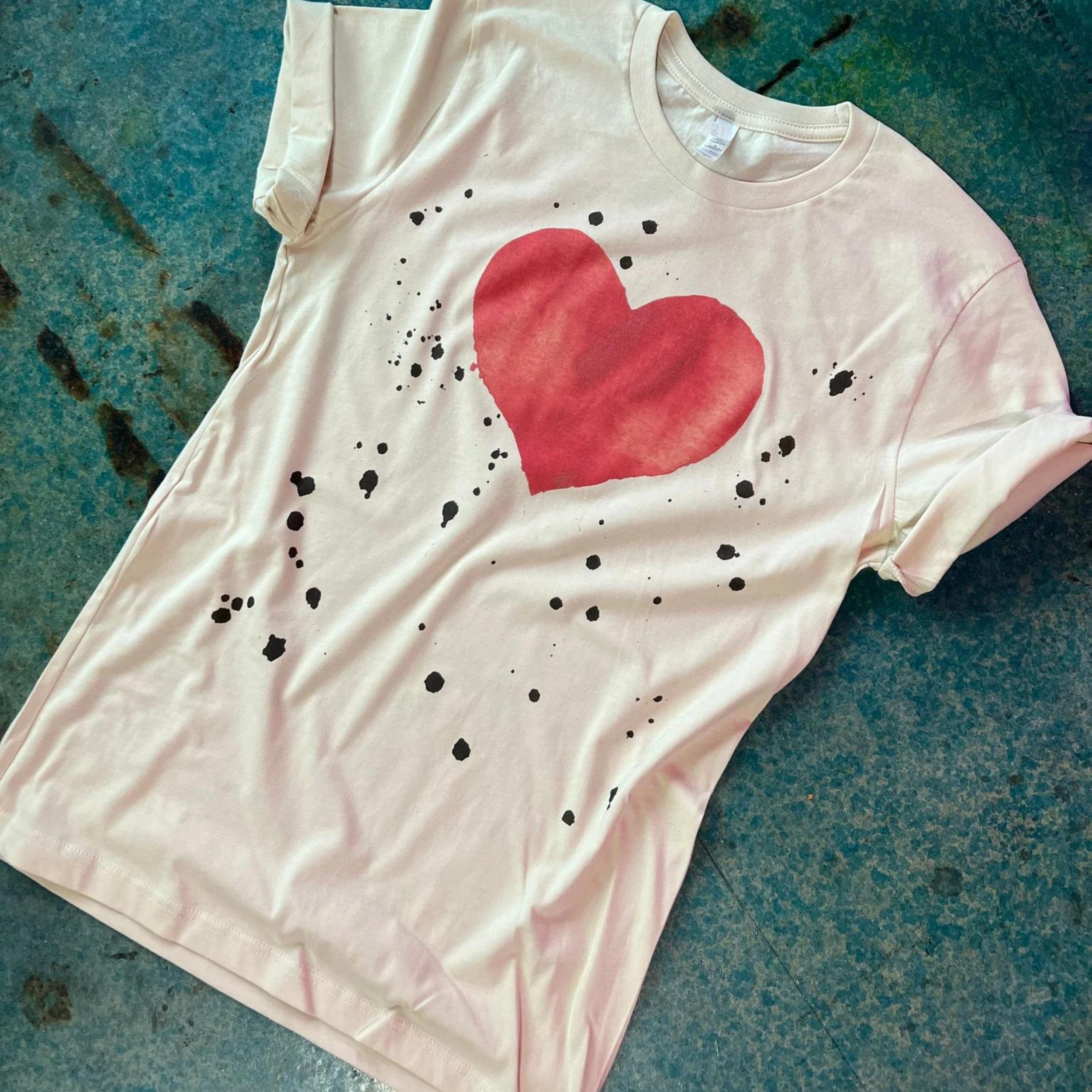 This cream tee is shown as a flatlay on a turquoise background. The graphic is a red heart with black paint splattered randomly on the front. This tee is also pictured with rolled sleeves. 