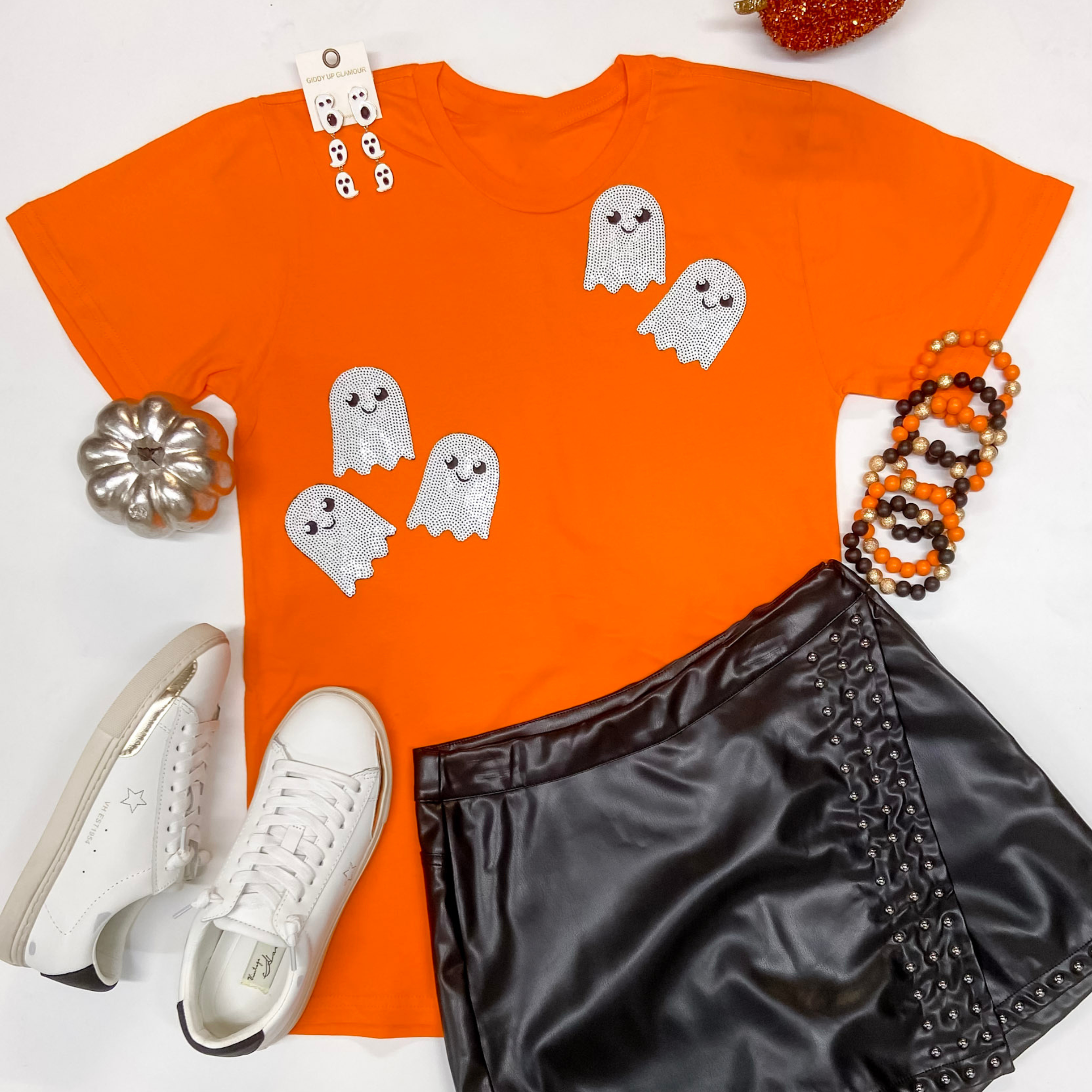 An orange short sleeve tee shirt with sequin ghosts pictured on a white background. This tee is pictured with white sneakers, a leather skort, and halloween jewelry.