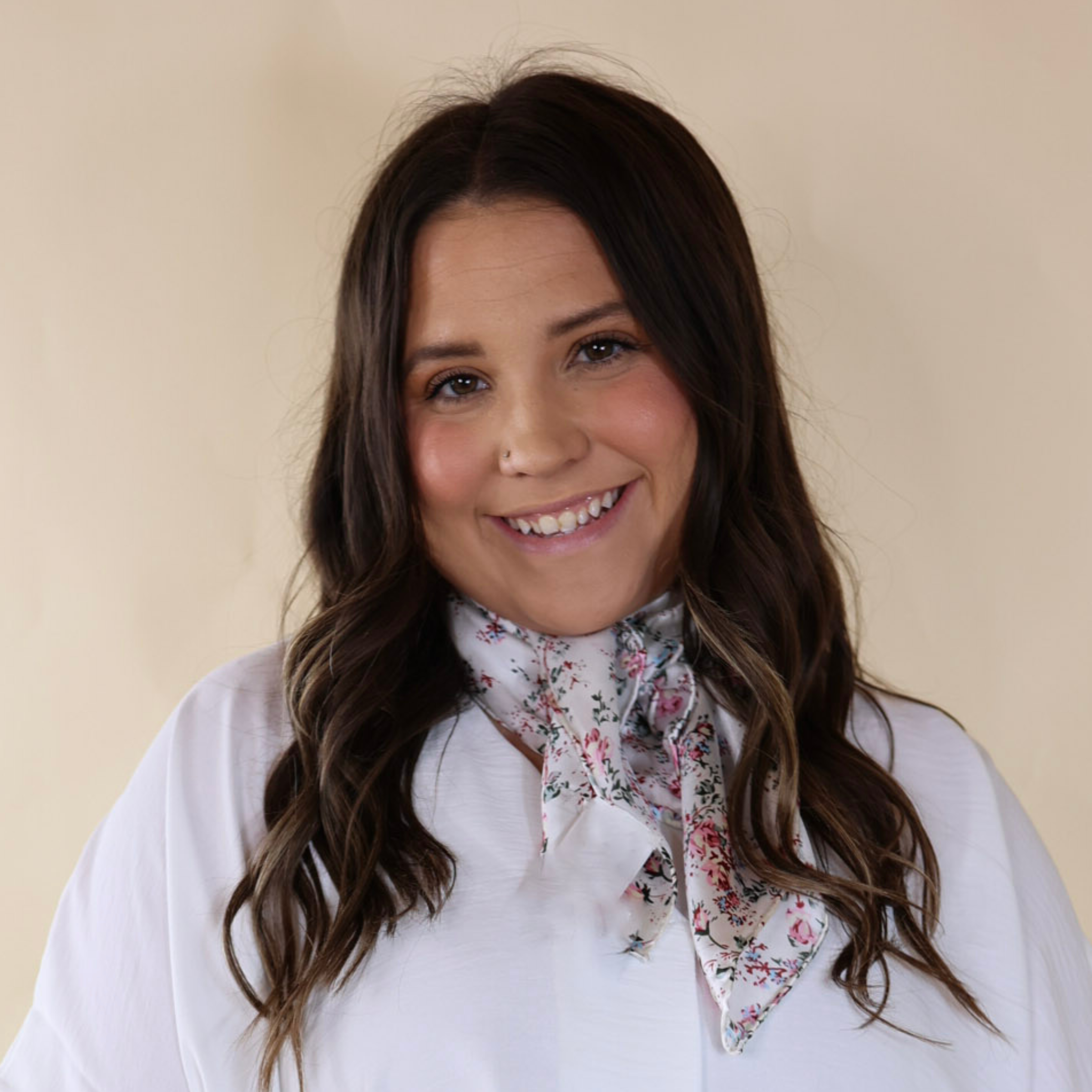 Brunette model wearing a white, off the shoulder top with a white scarf with a floral print tied around her neck. Model is pictured in front of a beige background. 