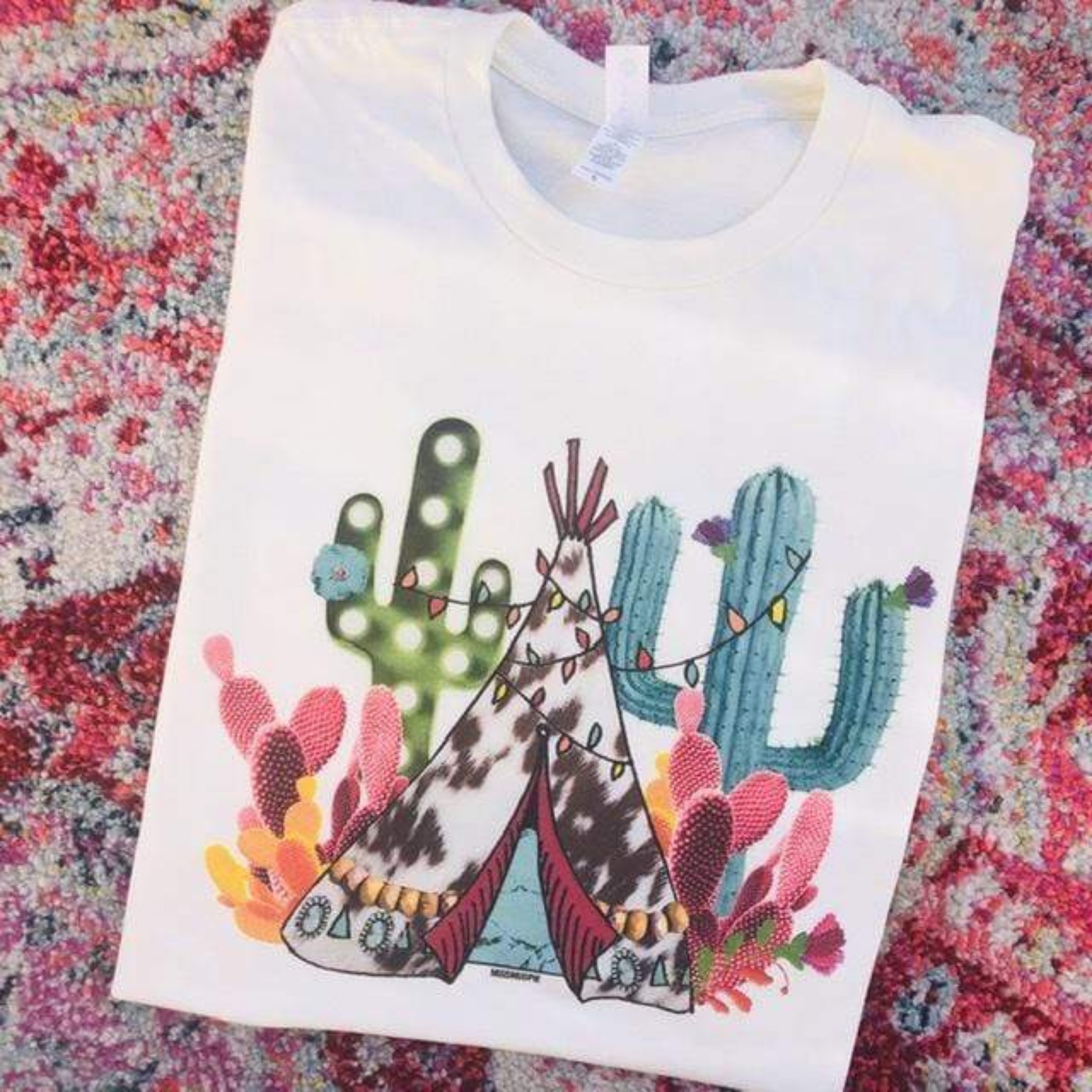 This white short sleeve crew neck tee with saguaro cactus and prickly pear cactus with Christmas lights and a cow print teepee. It is shown in this picture as a flat lay. 