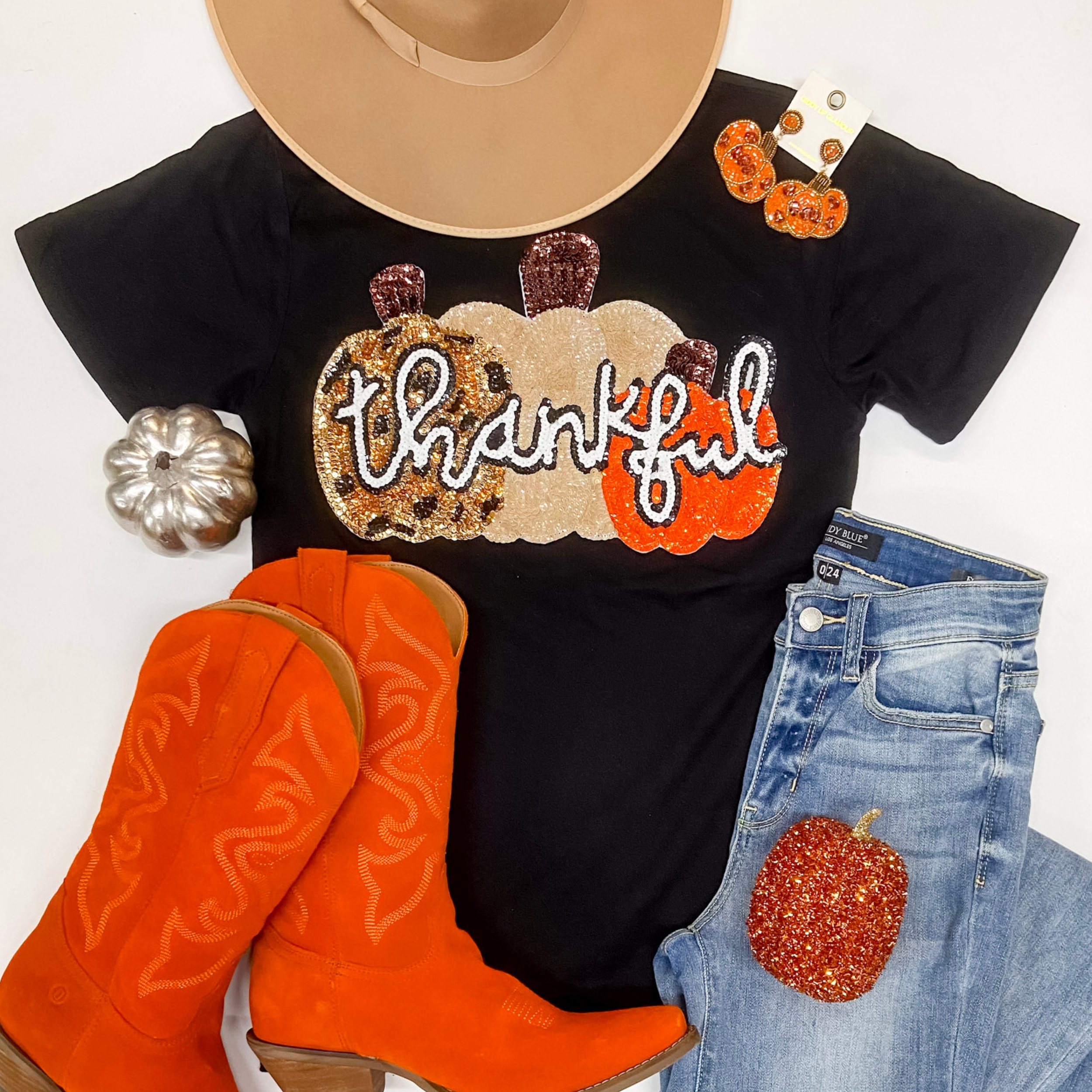 A black short sleeve graphic tee with a sequin patch that says "thankful" with pumpkins. This shirt is pictured on a white background with orange boots, light wash jeans, pumpkin earrings, and a tan hat.