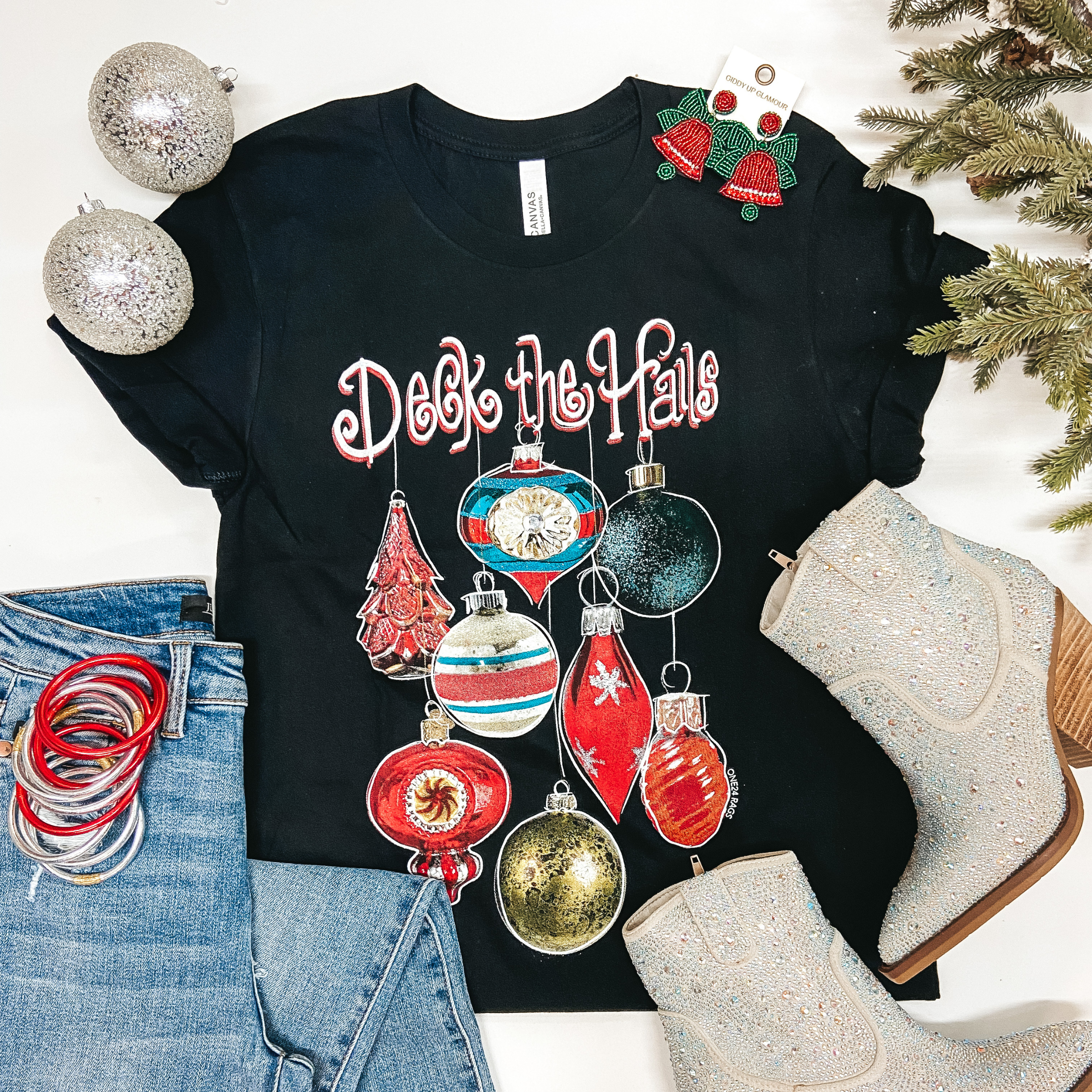 A solid black graphic tee with ornaments on the front and the phrase "Deck the Halls" written across the front. Pictured on a white background with silver booties, red earrings, silver and red bangles, and light wash jeans.