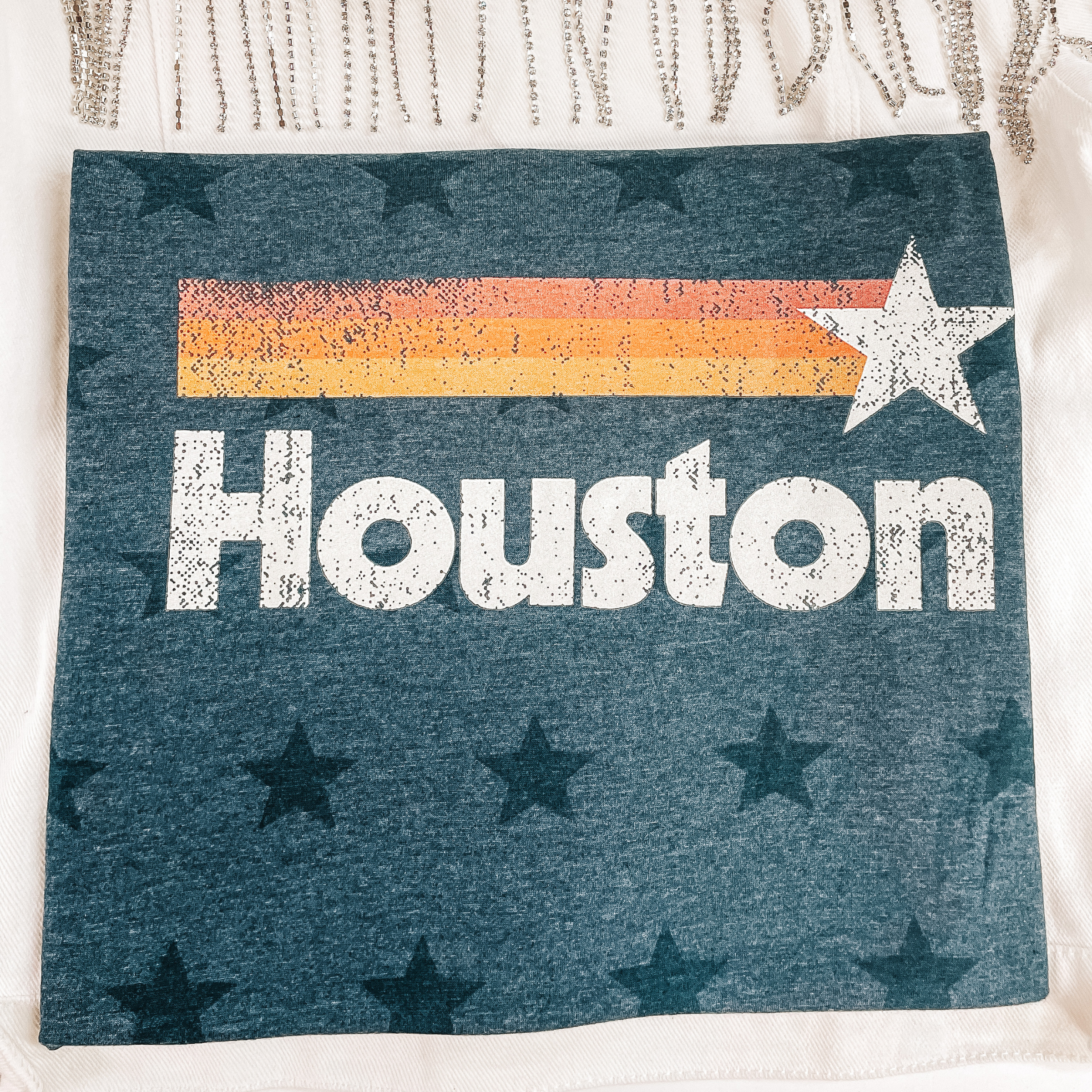 A folded tee shirt that is heather navy blue with a star print. The tee shirt has a graphic of a shooting star that says "Houston" in white letters underneath. Pictured on a white background with a white denim jacket. with crystal fringe.