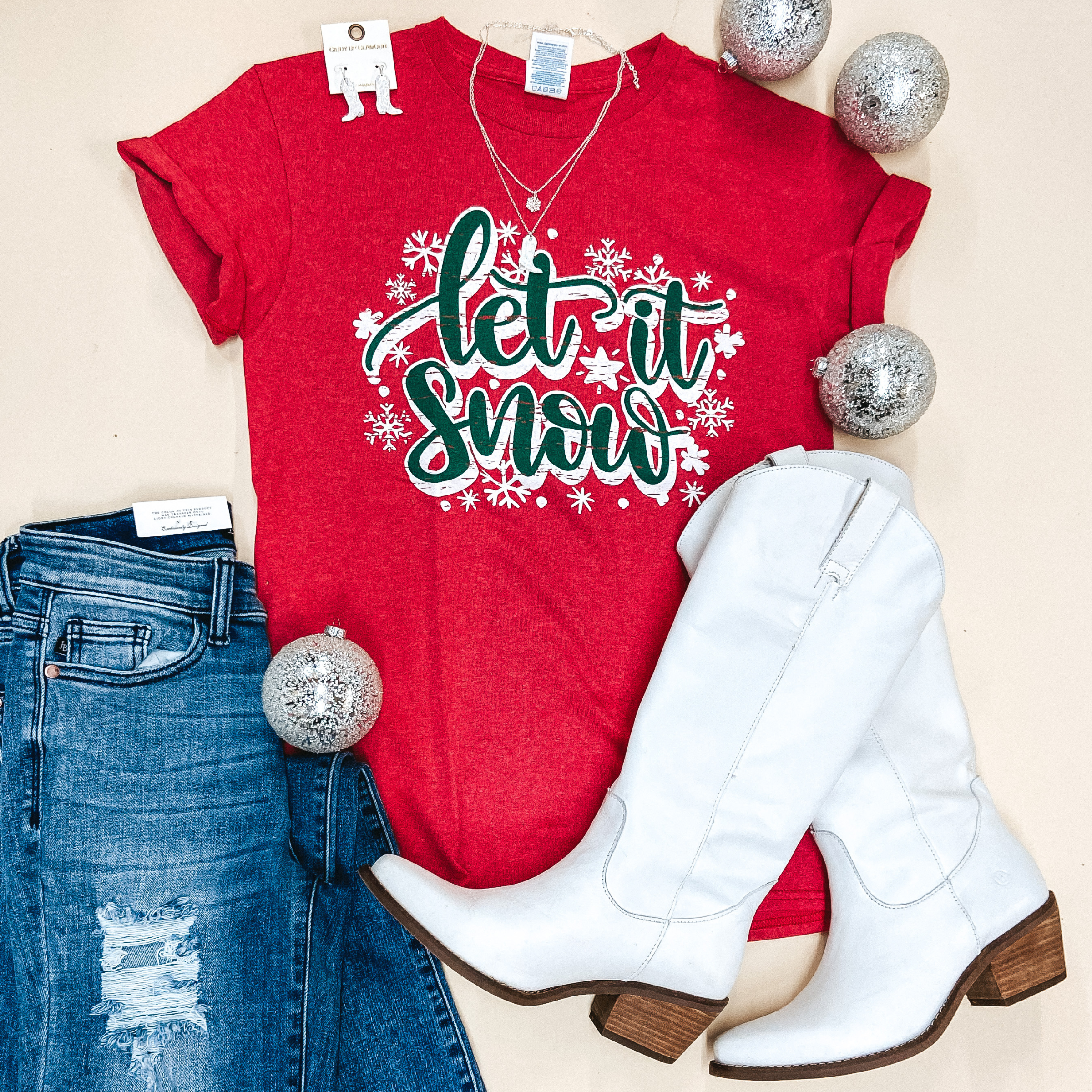 A red short sleeve tee shirt with a crew neckline and graphic that says "Let It Snow" in green lettering with snowflakes around. Pictured on a white background with distressed jeans, white jewelry, and white boots.