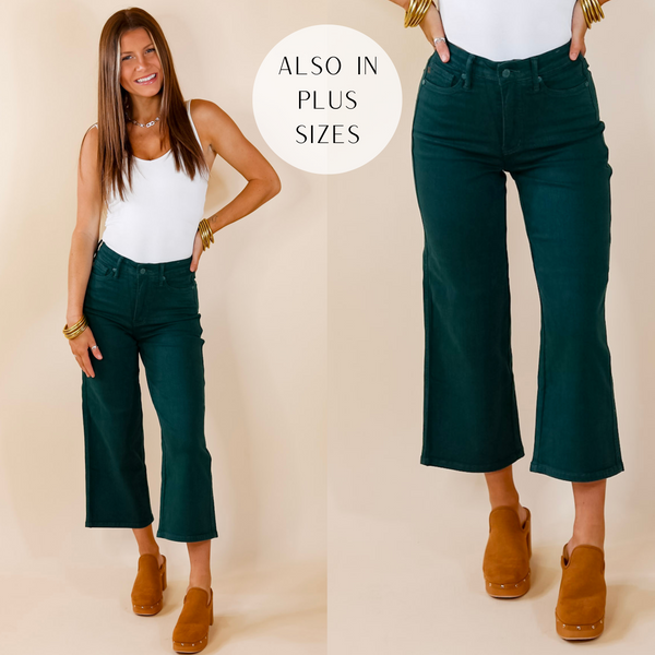 Model is wearing a pair of dark green cropped jeans. Model has them paired with a white bodysuit, tan clogs, and gold jewelry.