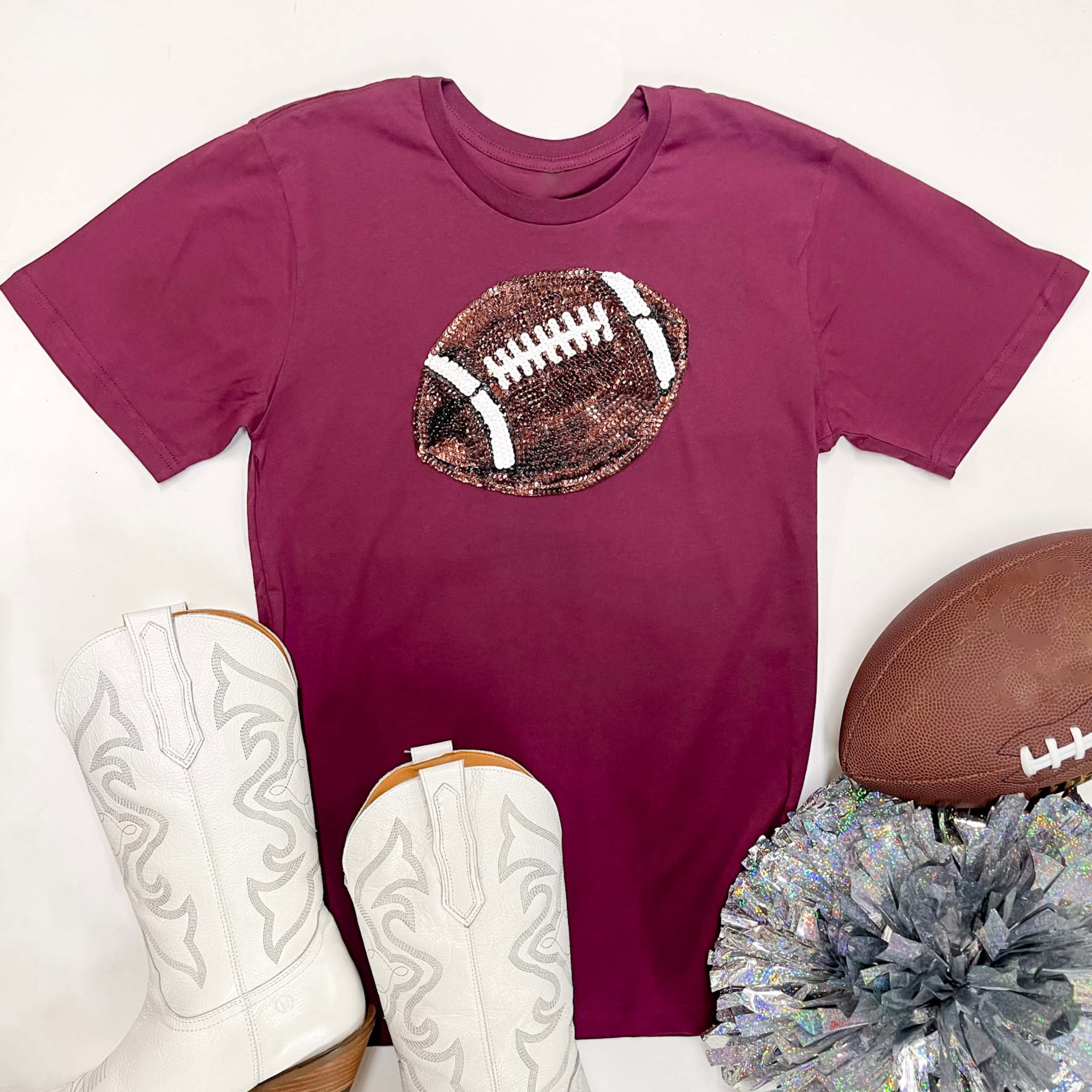 A maroon short sleeve graphic tee with a sequin football patch. This tee is paired with white boots, a silver pom pom, and a brown football.