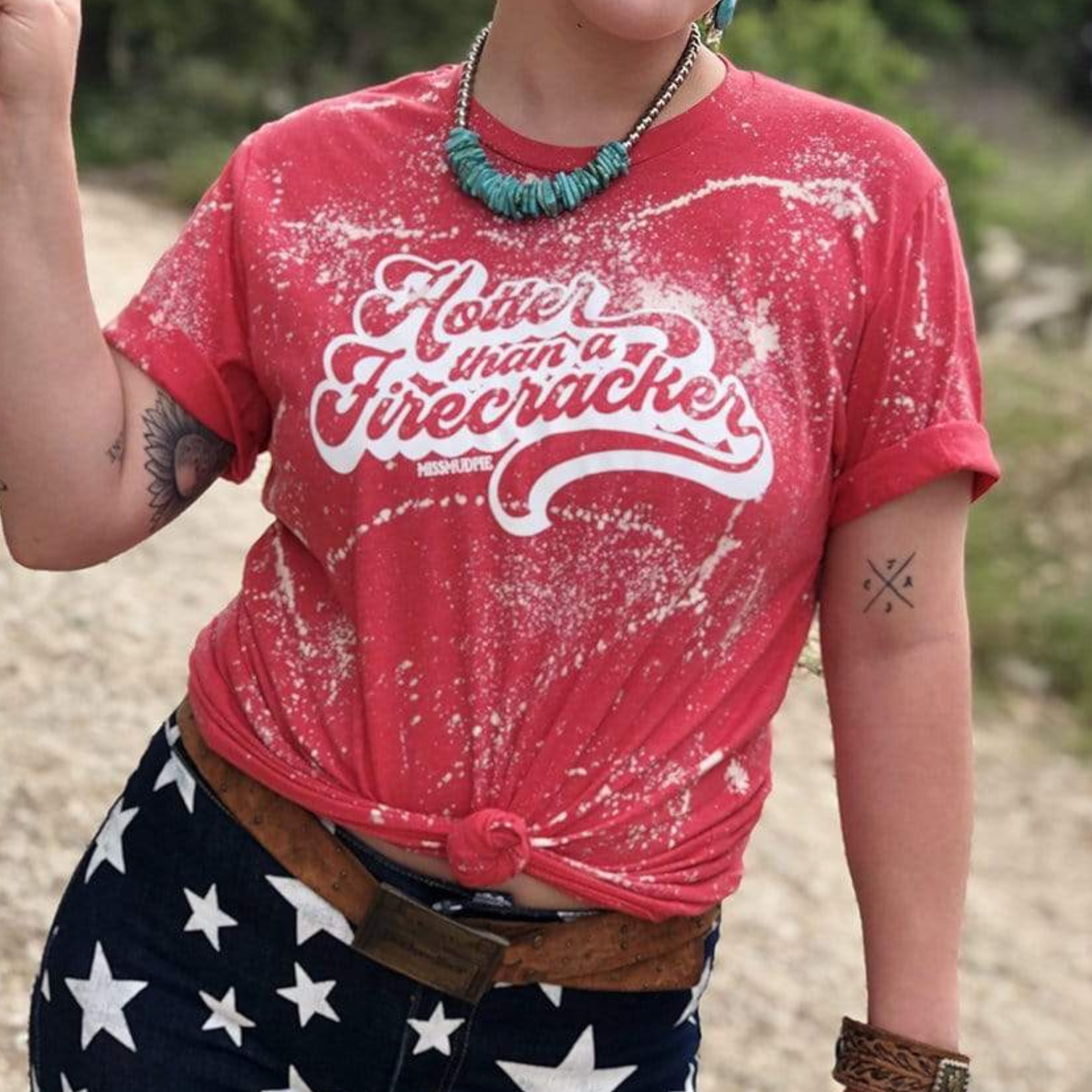  red short sleeve tee with a splatter paint design with the words "Hotter than a firecracker" in cursive in the center. Item is pictured on a beige and green background