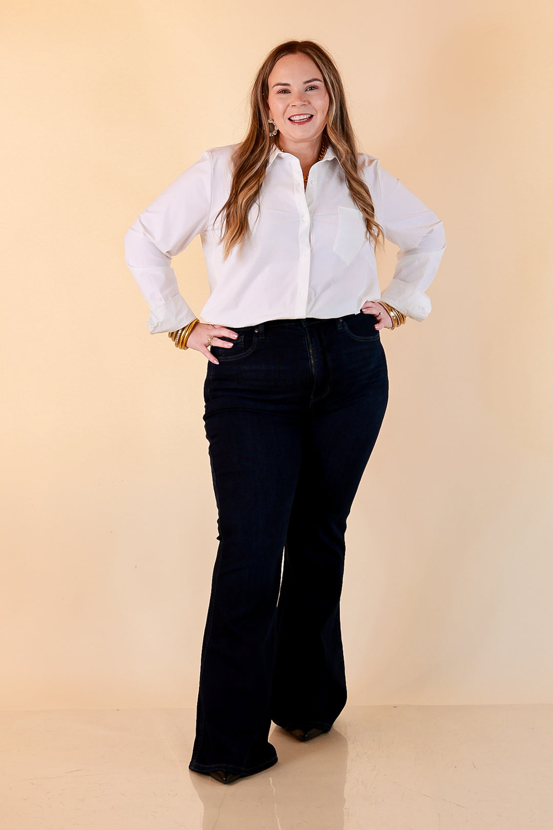 A woman in a white button-up shirt and dark wash Judy Blue tummy control flare jeans with gold bangles and black pointed-toe heels